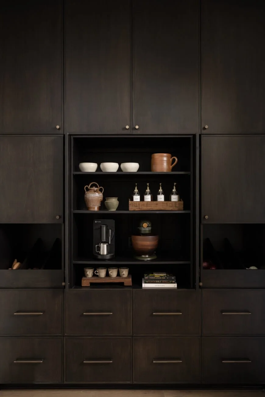 Built-in black kitchen cabinet with open shelves holding cups, bowls, a coffee maker, and books.