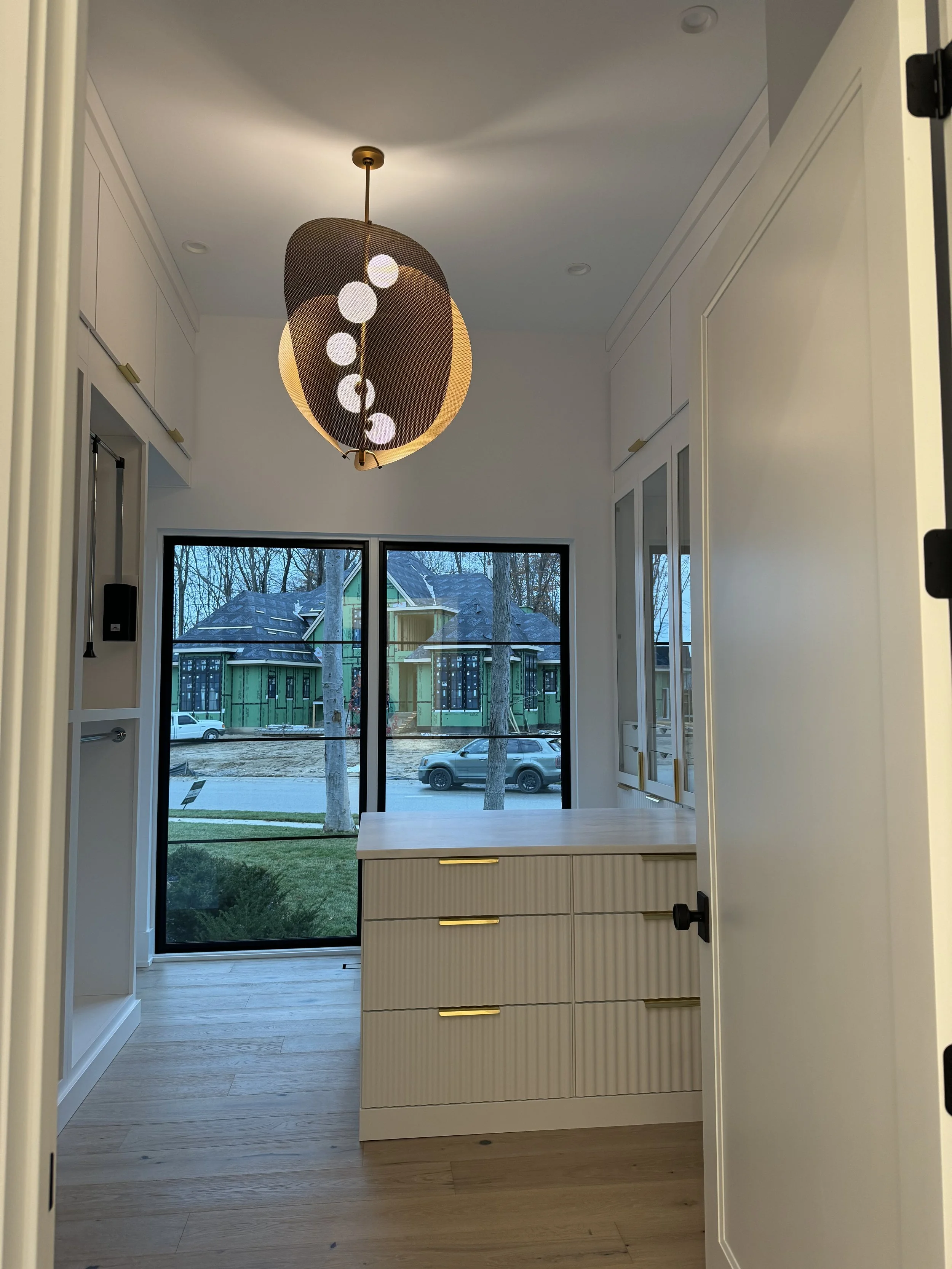 View through a doorway into a modern kitchen with a white kitchen island, a large window showing trees and neighboring houses, and a pendant light fixture hanging from the ceiling.