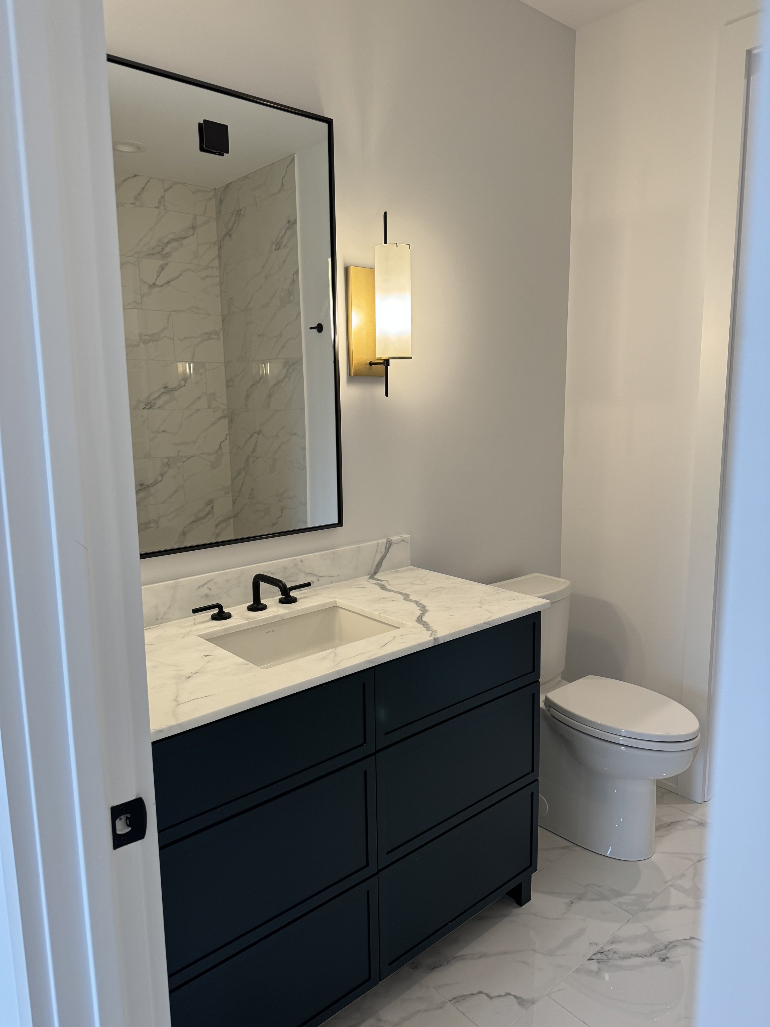 Modern bathroom with a dark blue vanity, marble countertop, large mirror, wall-mounted light fixture, marble-tiled shower area, and a white toilet.
