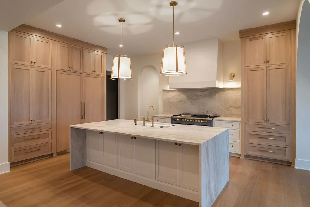 Modern kitchen with light wood cabinets, marble island and backsplash, gold fixtures, and pendant lights.