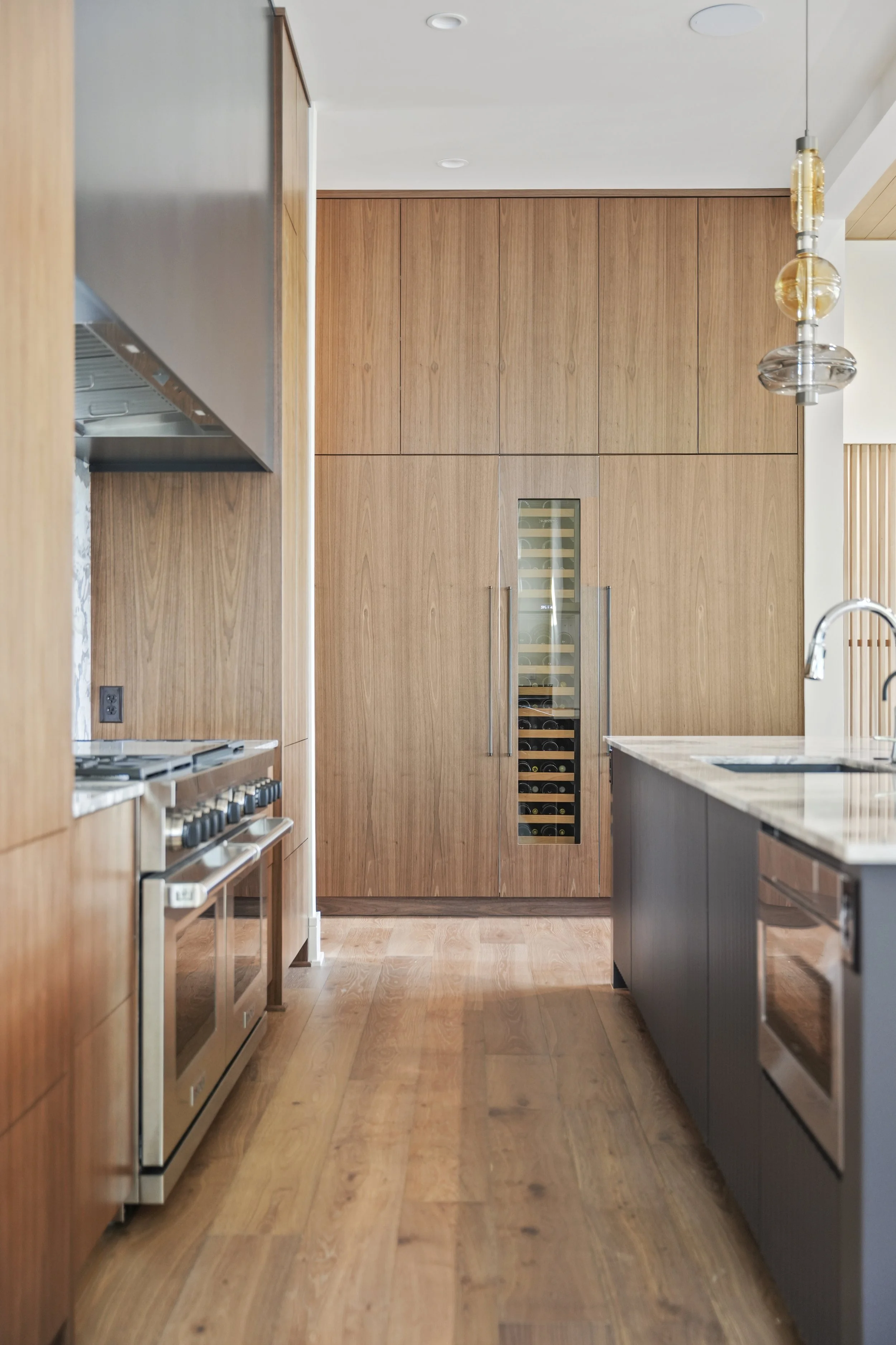 Modern kitchen with wooden cabinetry, stainless steel appliances, and a wine fridge built into the cabinetry. Pendant lights hang from the ceiling over a marble countertop island, with wood flooring throughout.