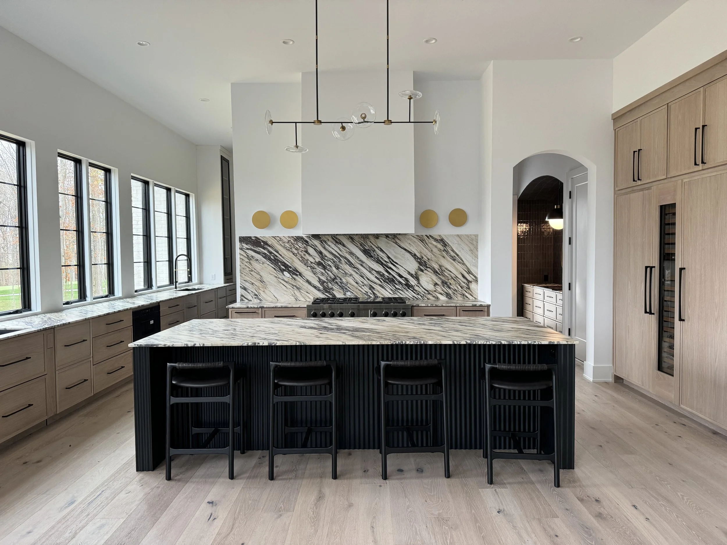 Modern kitchen with a white countertop and backsplash, black stove, light wood cabinets, black pendant chairs, large windows, and a contemporary chandelier.