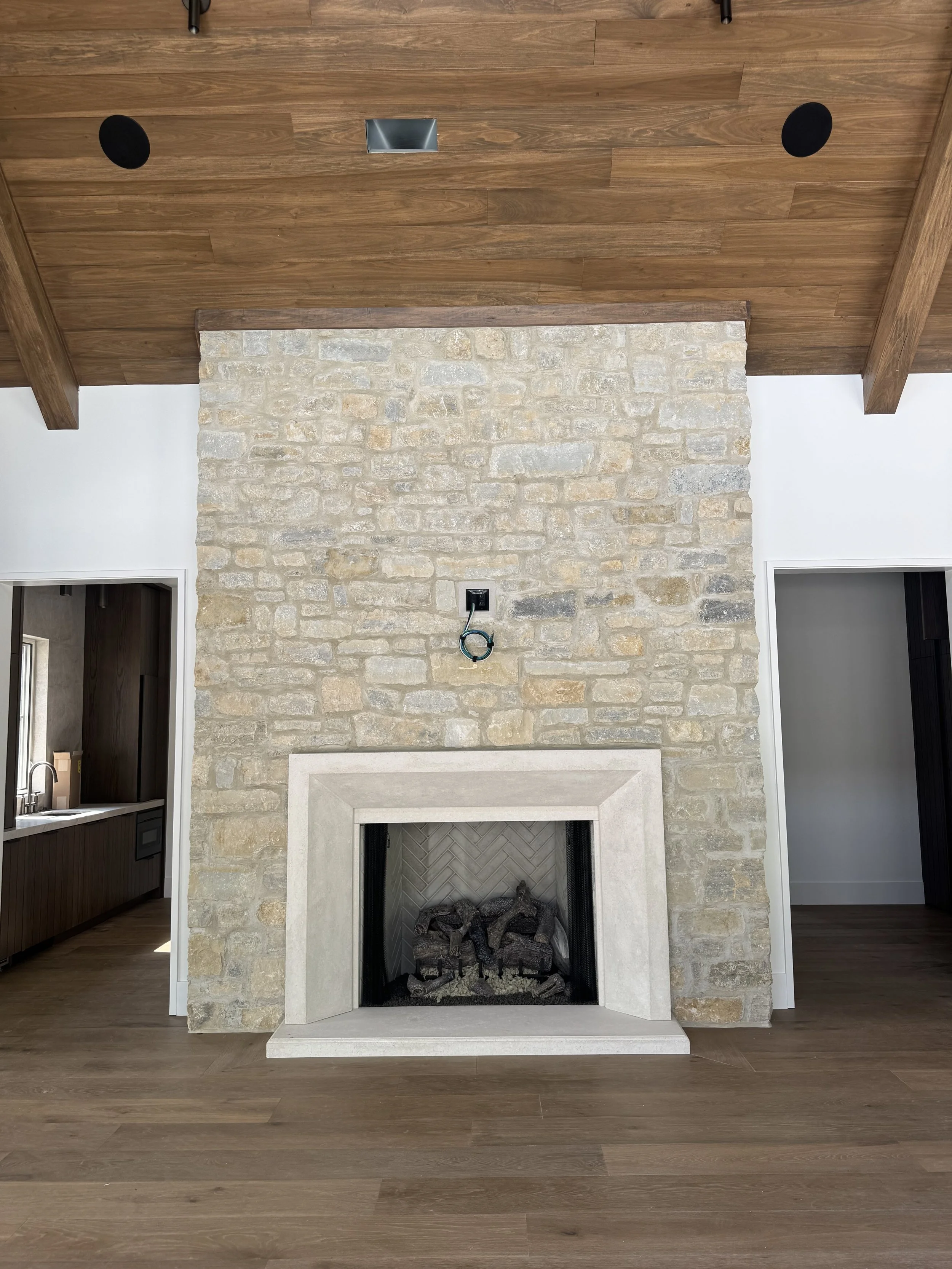 A stone fireplace with a white mantel in a living room, with a wood-paneled ceiling and hardwood floors.