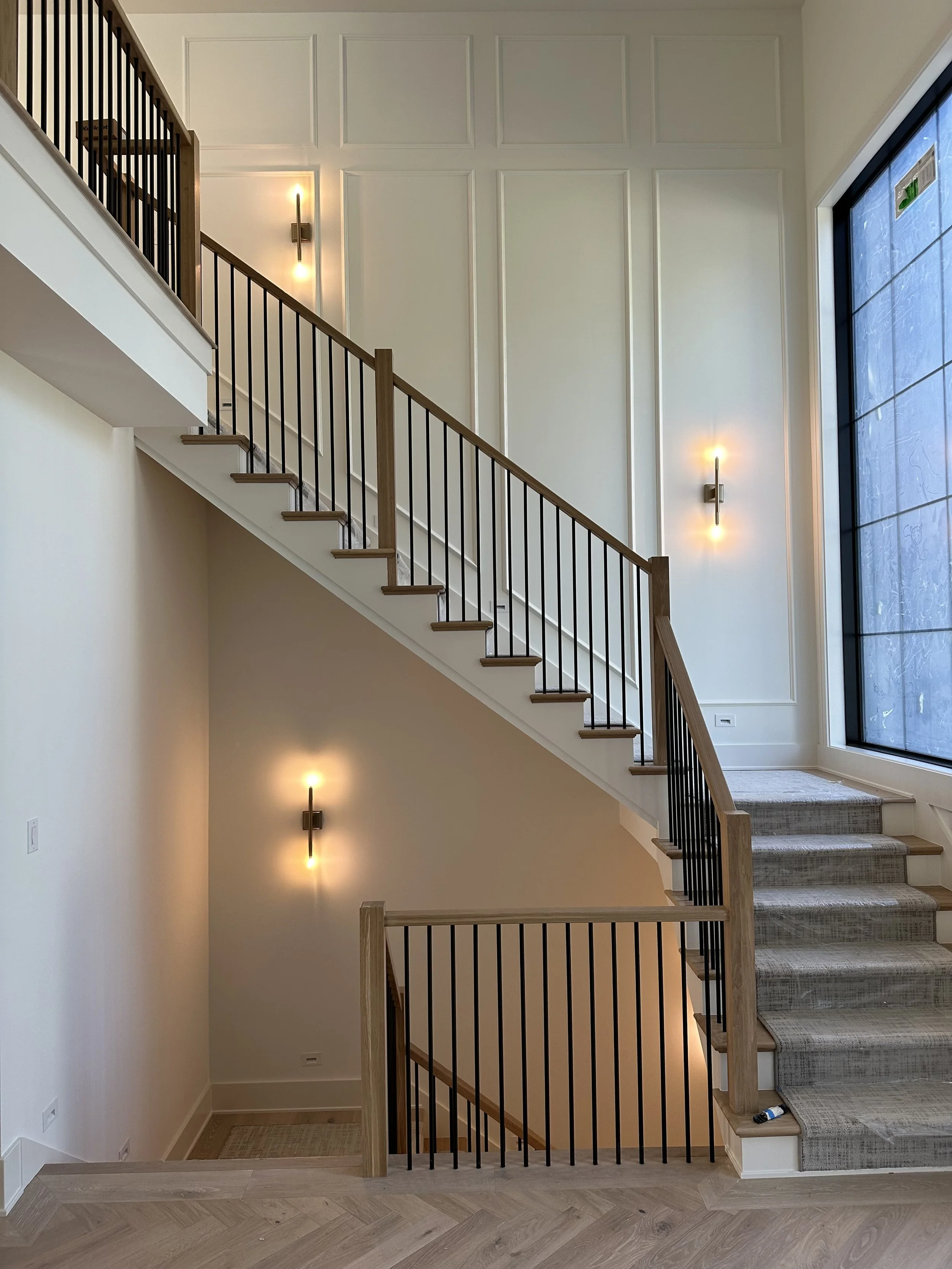 Interior view of a modern staircase with wood steps, black metal railings, and beige carpeting on the stairs. The wall features decorative paneling with built-in warm lighting fixtures. Large window on the right side allows natural light into the spa