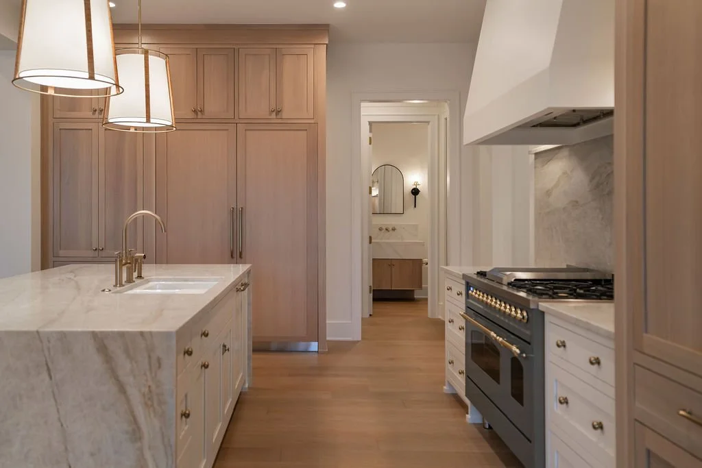 Modern kitchen with light wood cabinets, marble countertops, gold hardware, a blue stove, and pendant lighting. A doorway leads to a nearby bathroom with a mirror and vanity.
