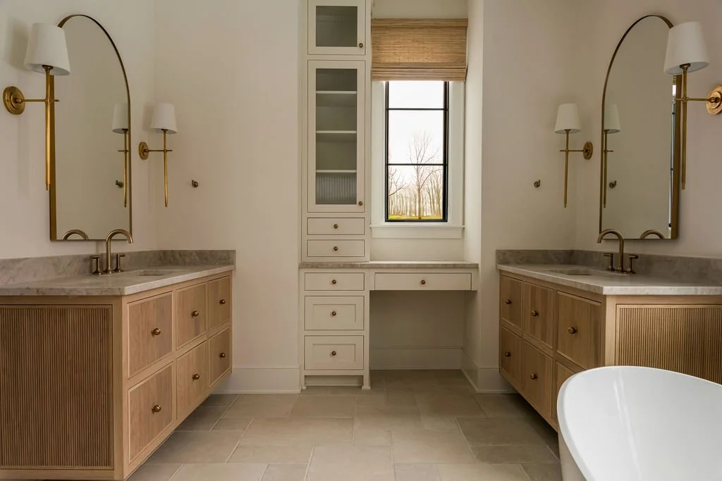 A bathroom with two wooden vanities, each with a marble countertop and a brass faucet, mirrors, and wall-mounted sconces. There is a window with a bamboo shade providing natural light, and built-in white cabinets and drawers between the vanities.