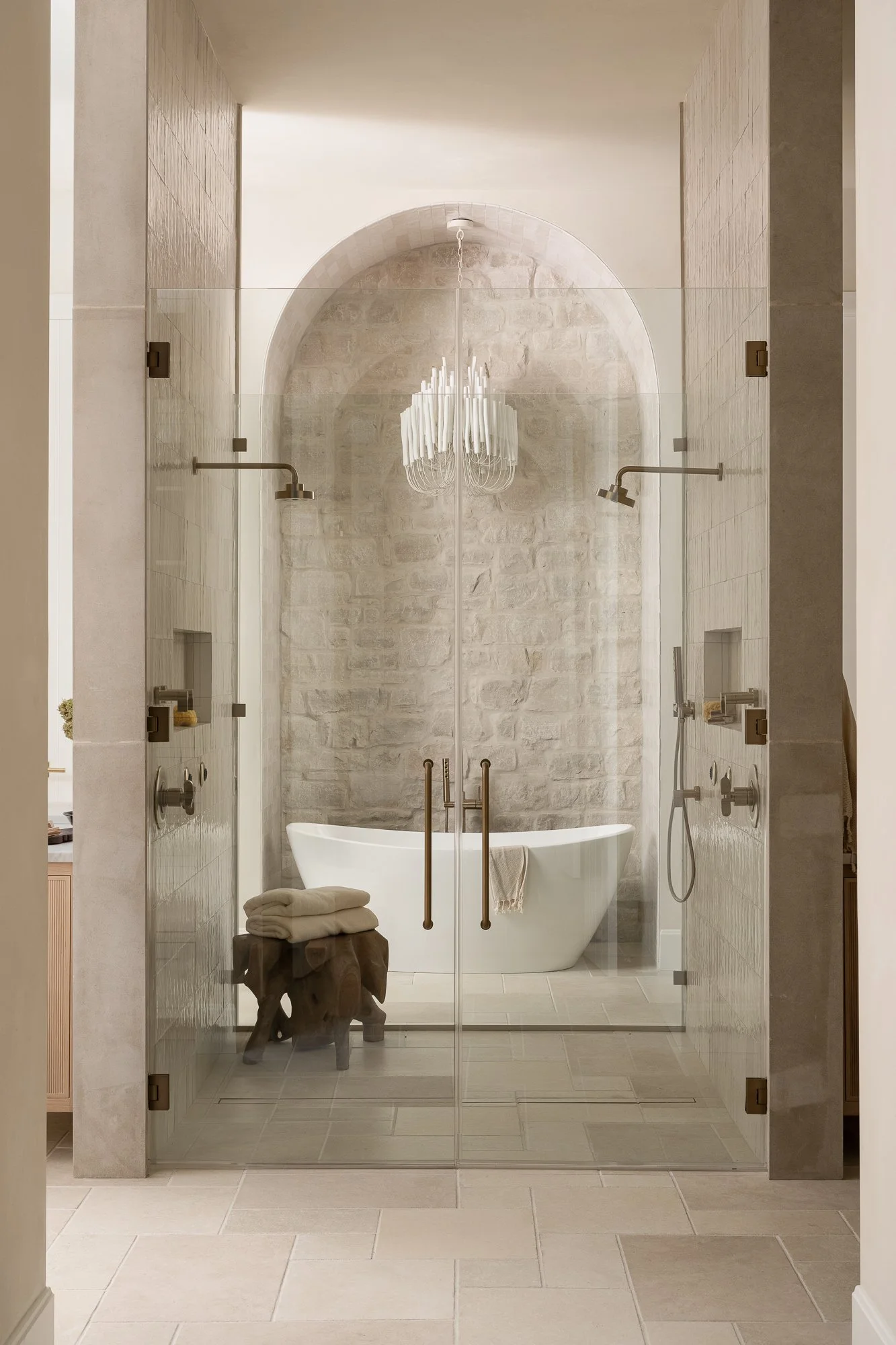 Modern bathroom with a standalone bathtub, glass shower enclosure, stone wall, chandelier, and neutral-toned tiles.