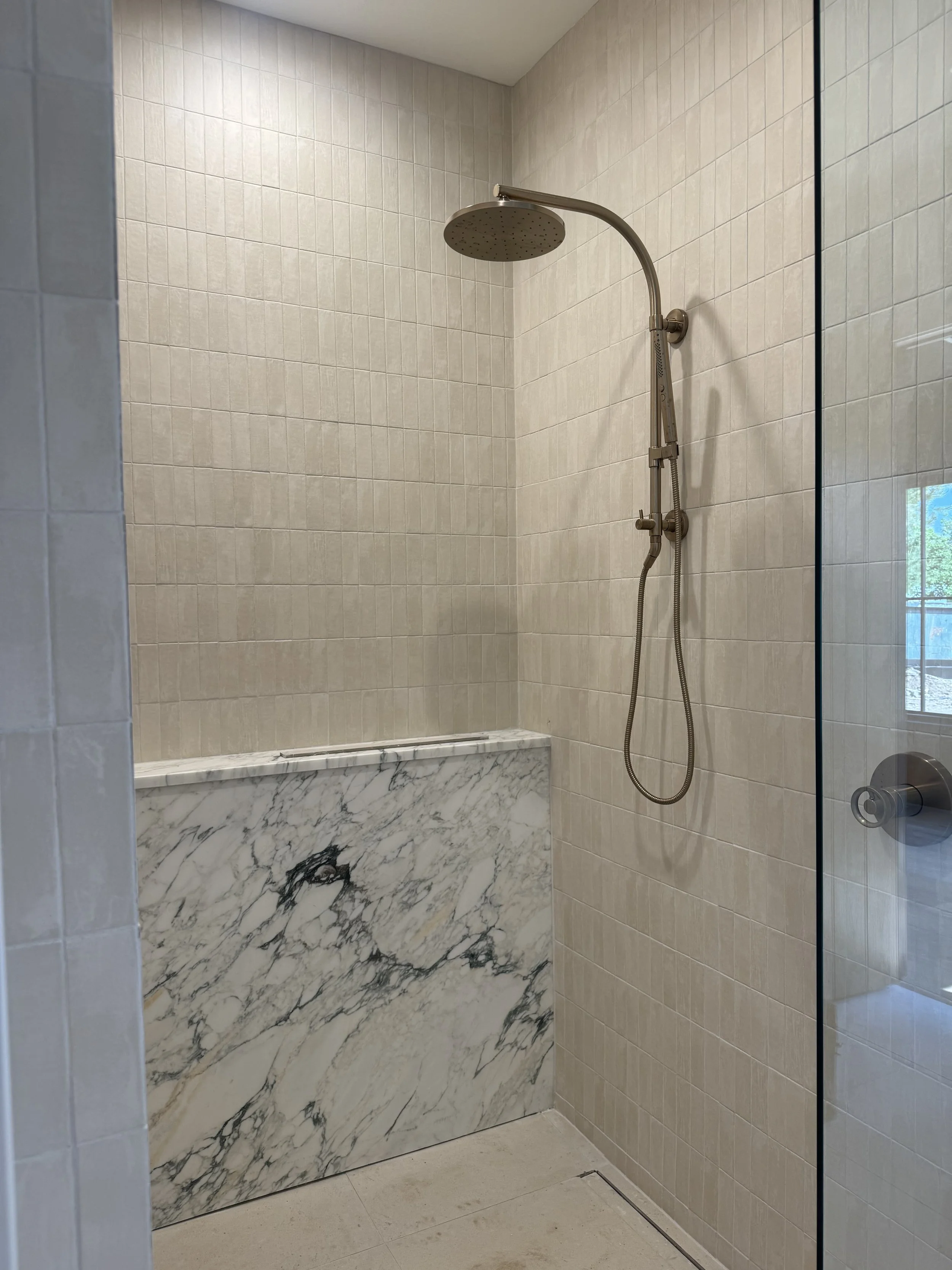 A modern shower with beige tiled walls, a large round shower head, a handheld shower attachment, and a marble ledge at the base, inside a glass enclosure.