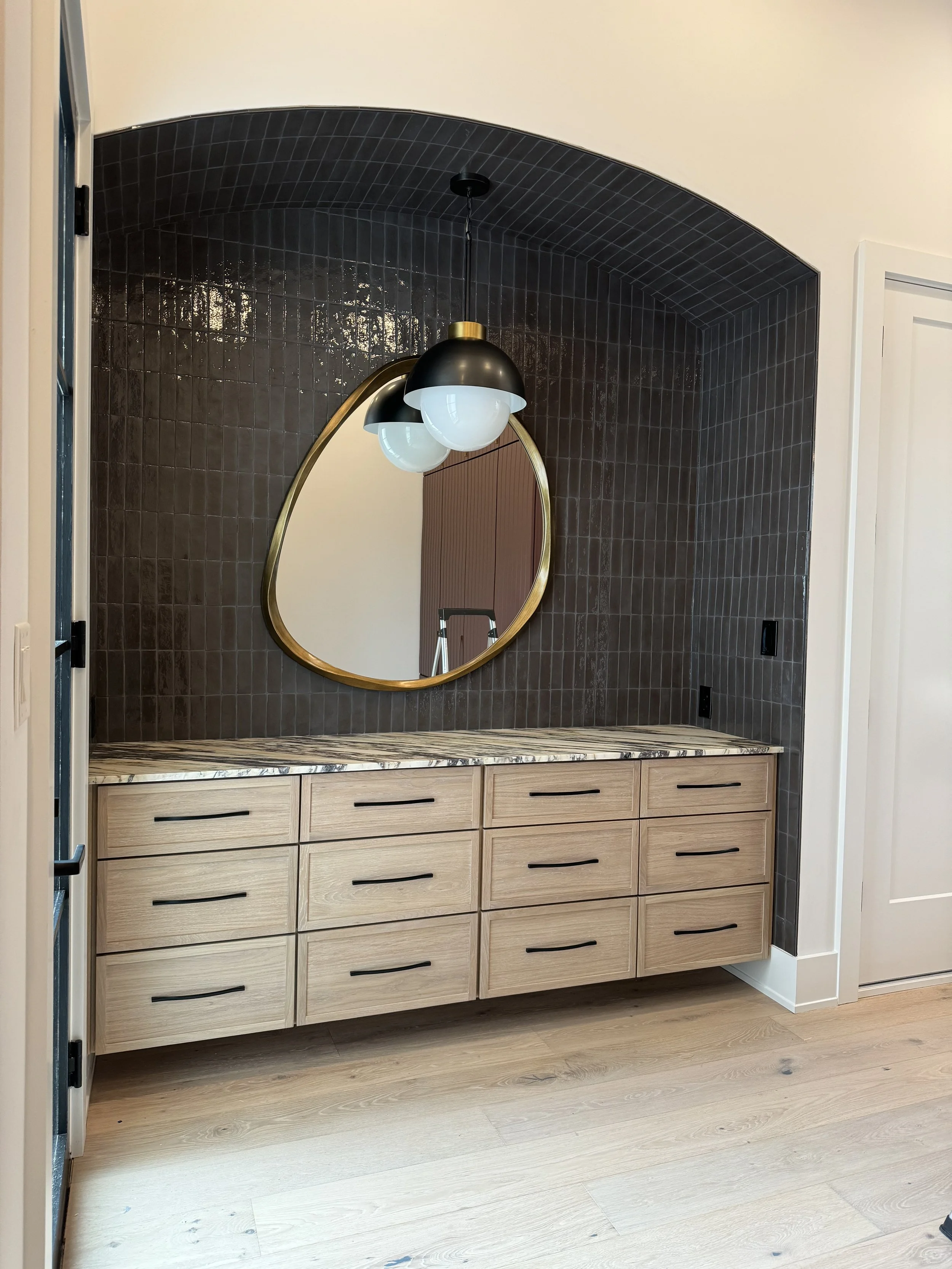 Modern bathroom vanity with wooden drawers, marble countertop, black and gold mirror, dark tiled wall, and hanging light fixtures.