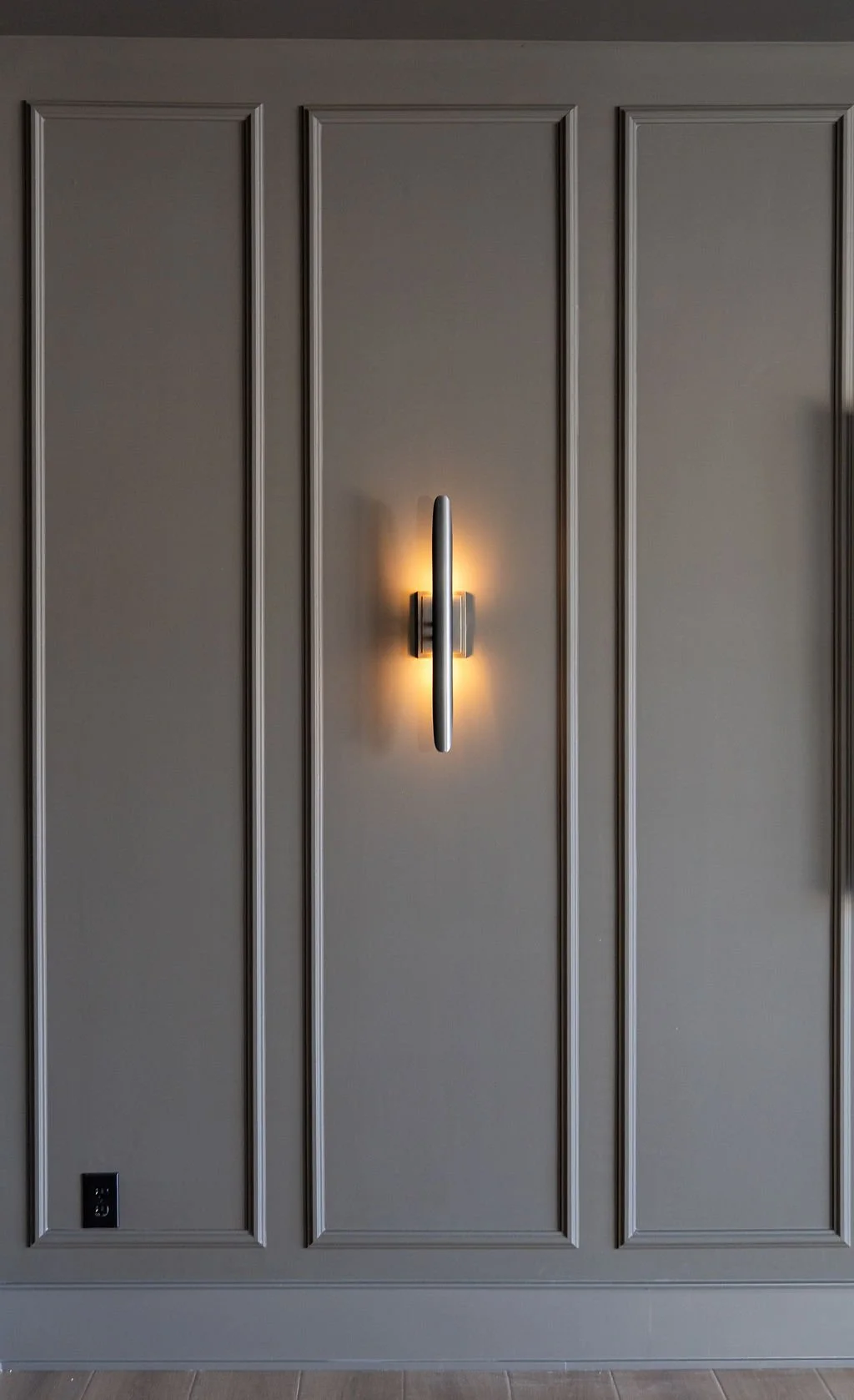 Gray wall with decorative molding and a modern wall sconce light fixture that is illuminated.