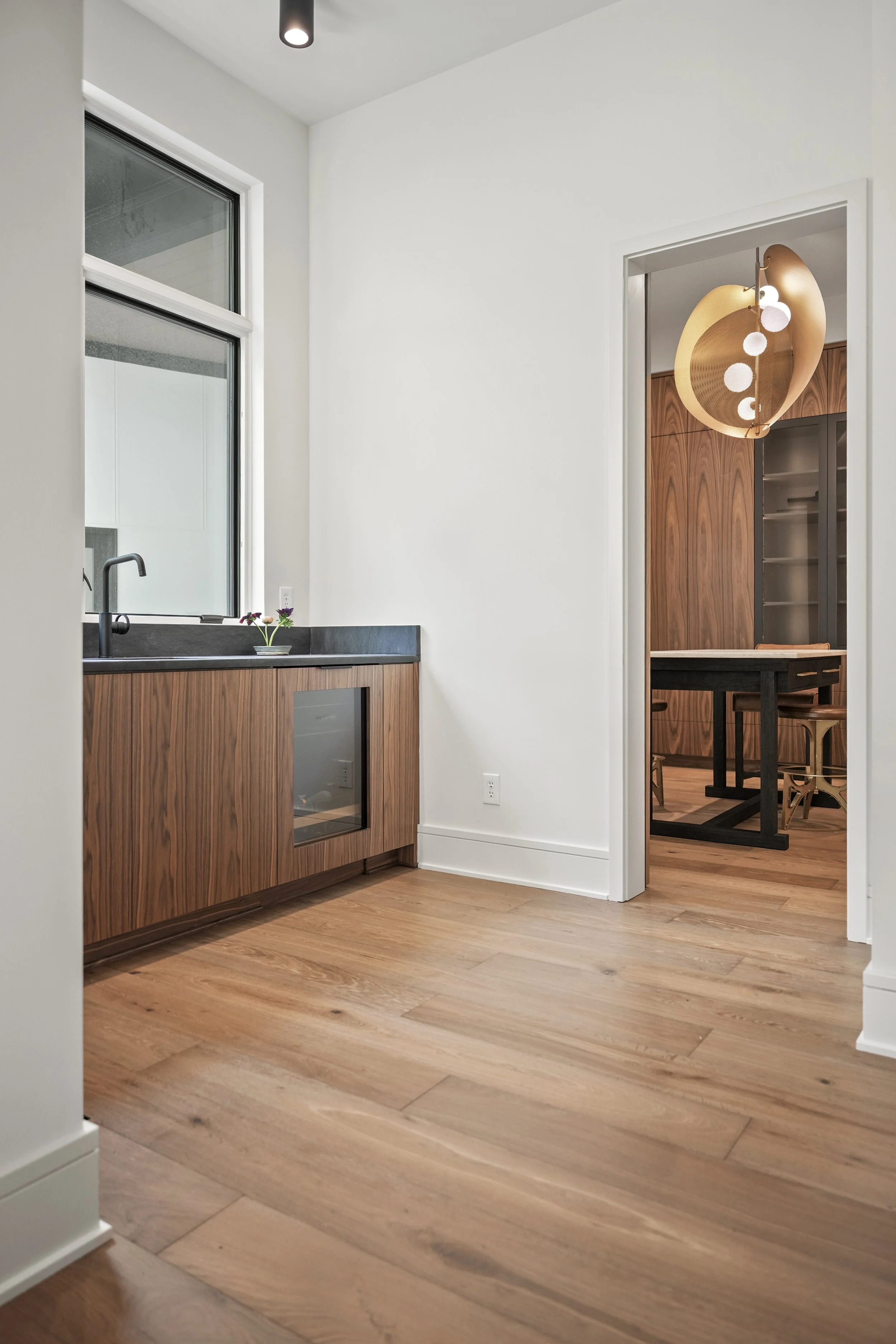 A modern kitchen with wooden cabinetry, a black countertop, large window, and a small plant; adjacent dining area with wood-paneled cabinets and a black dining table with wooden chairs, illuminated by a contemporary hanging light fixture.