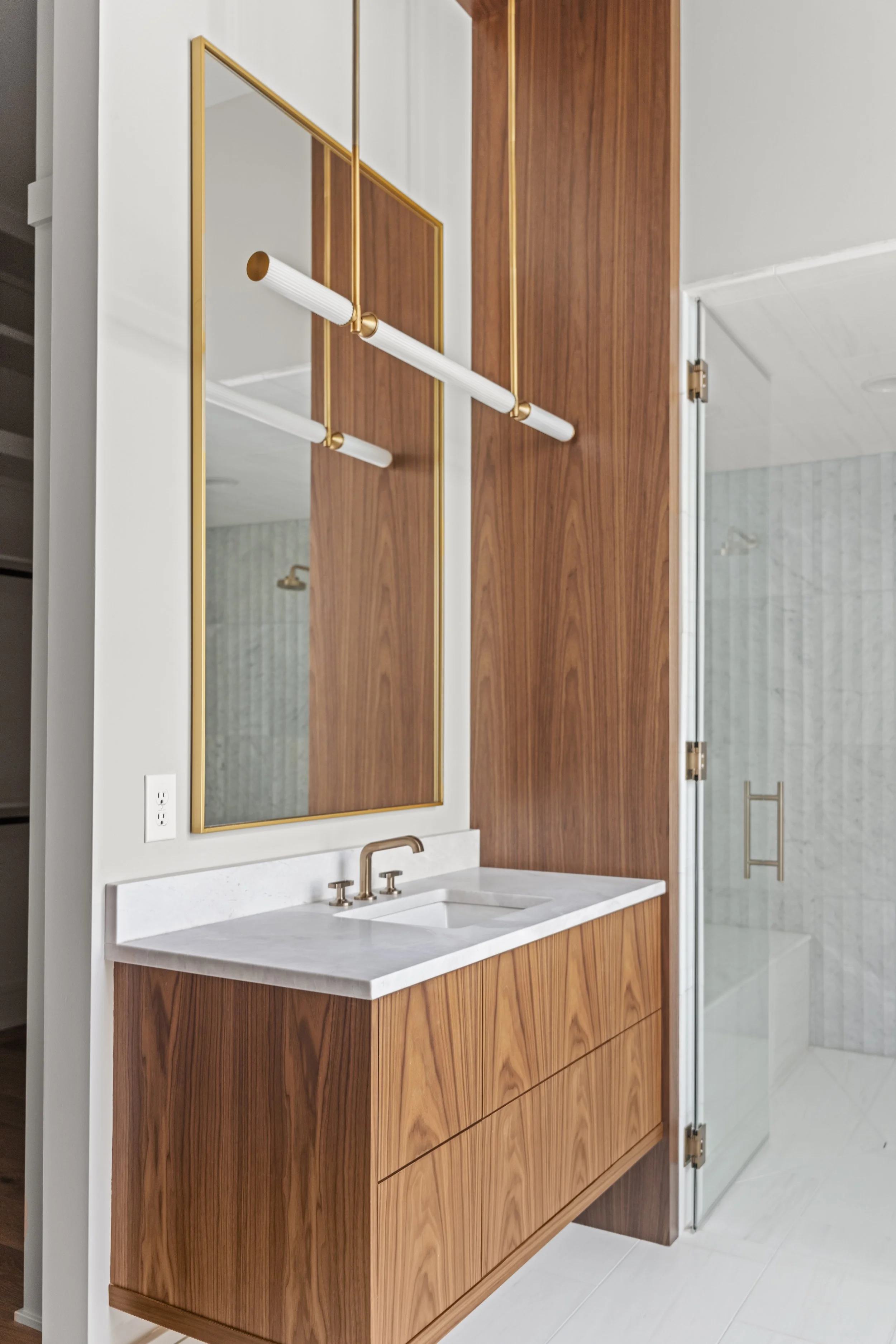 Modern bathroom vanity with wooden cabinet, white countertop, and gold fixtures, next to a glass shower door with a gold handle.