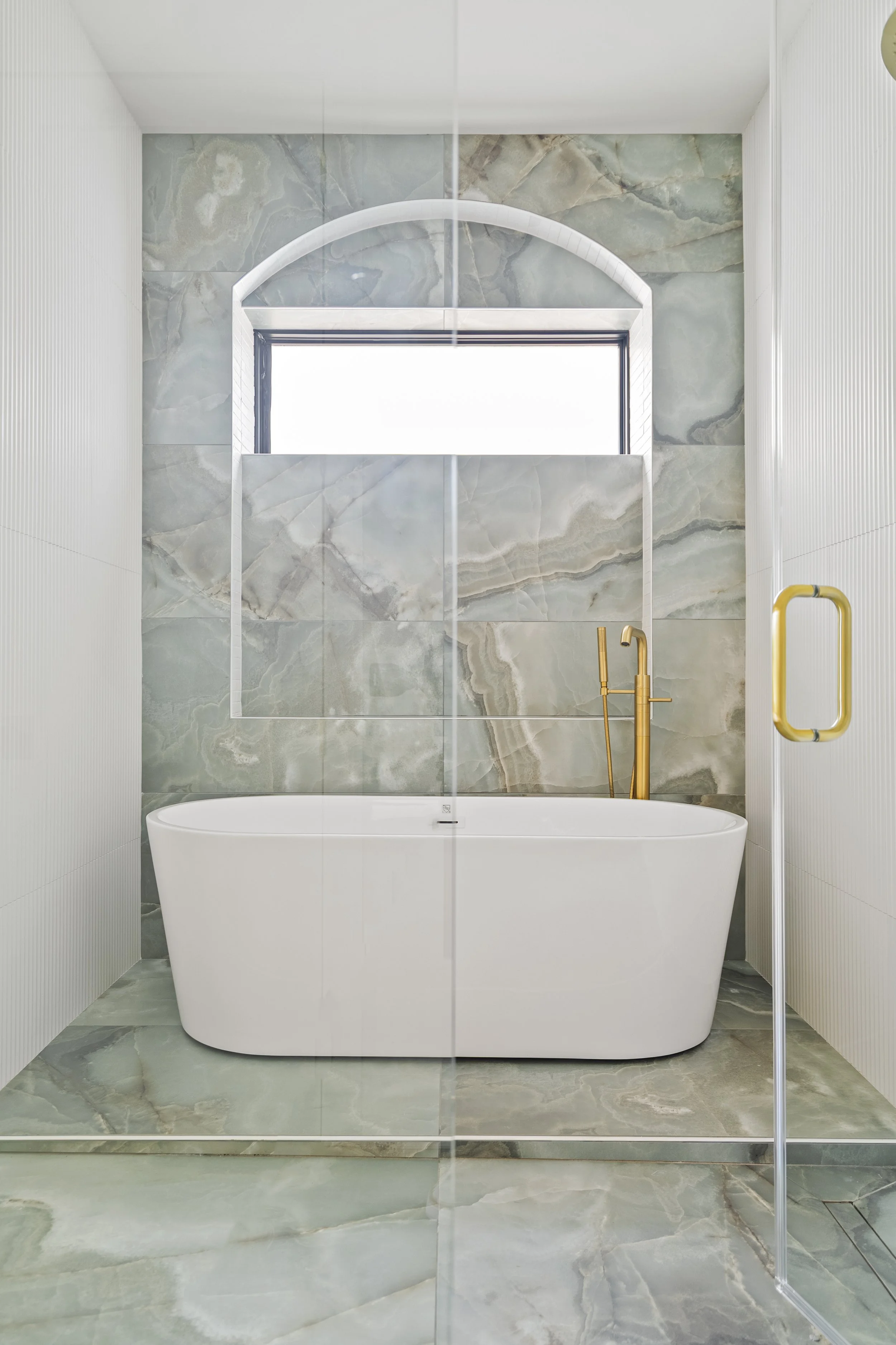 Modern bathroom with freestanding bathtub, marble walls, and a window. Golden fixtures and a glass door are also visible.