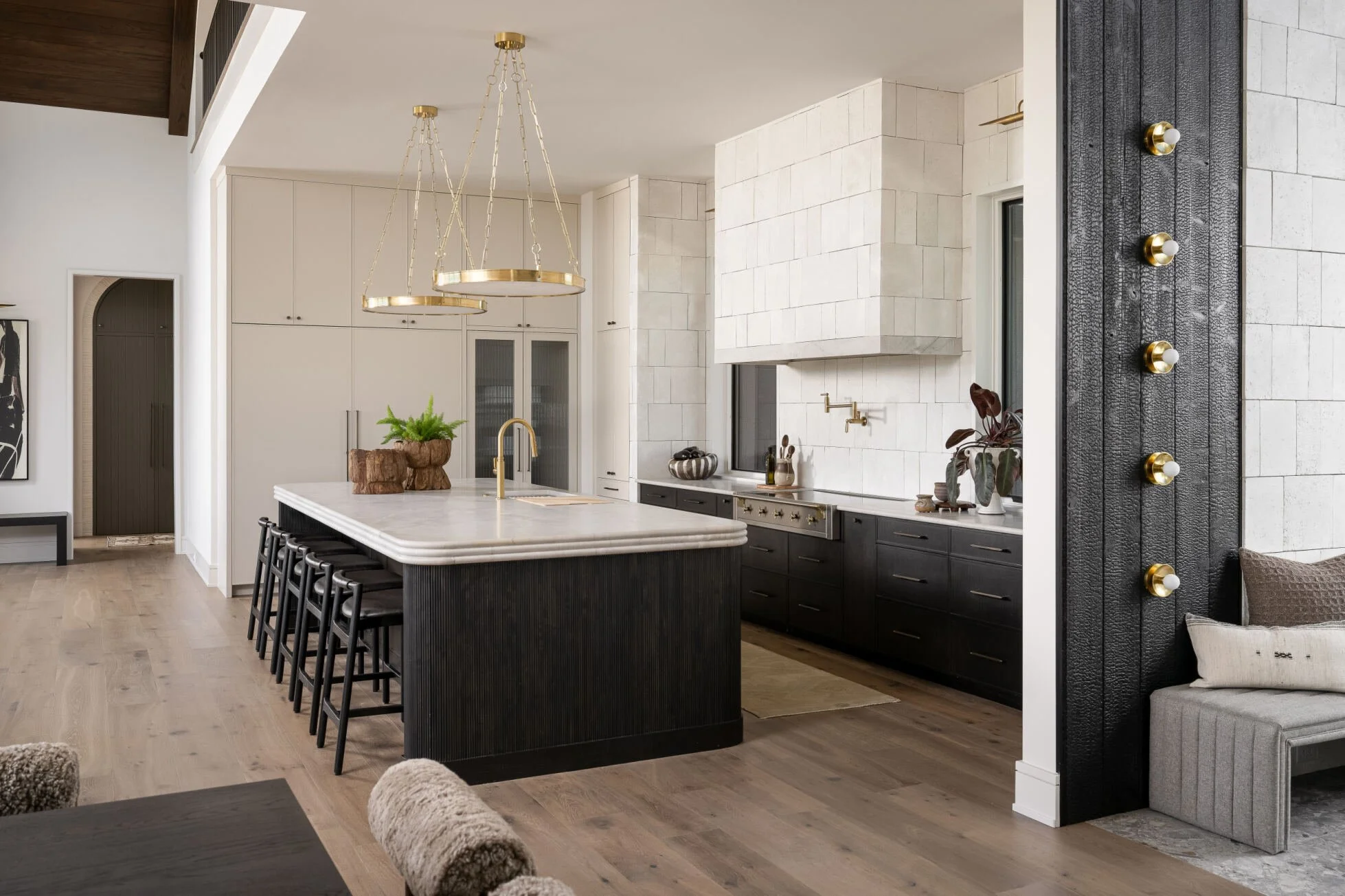 Modern kitchen with white and black cabinetry, a large kitchen island with bar stools, light wood floors, and a fireplace with gold faucet fixtures. Pendant lights hang above the island, and there are decorative plants on the counter.