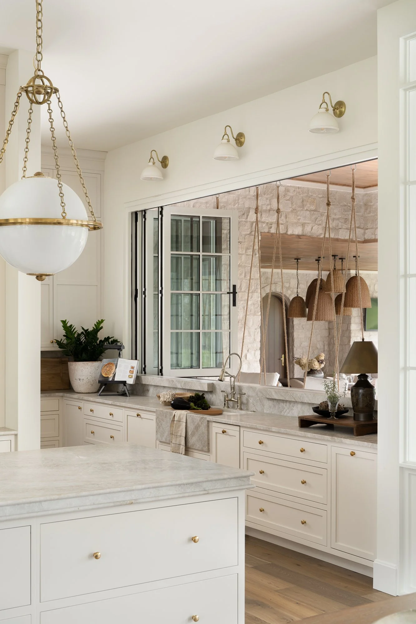 A bright, modern kitchen with white cabinets and a marble countertop. There are three wall-mounted light fixtures, a contemporary hanging light with chains, and a view into a living area with a stone wall, hanging woven lamps, and large windows.