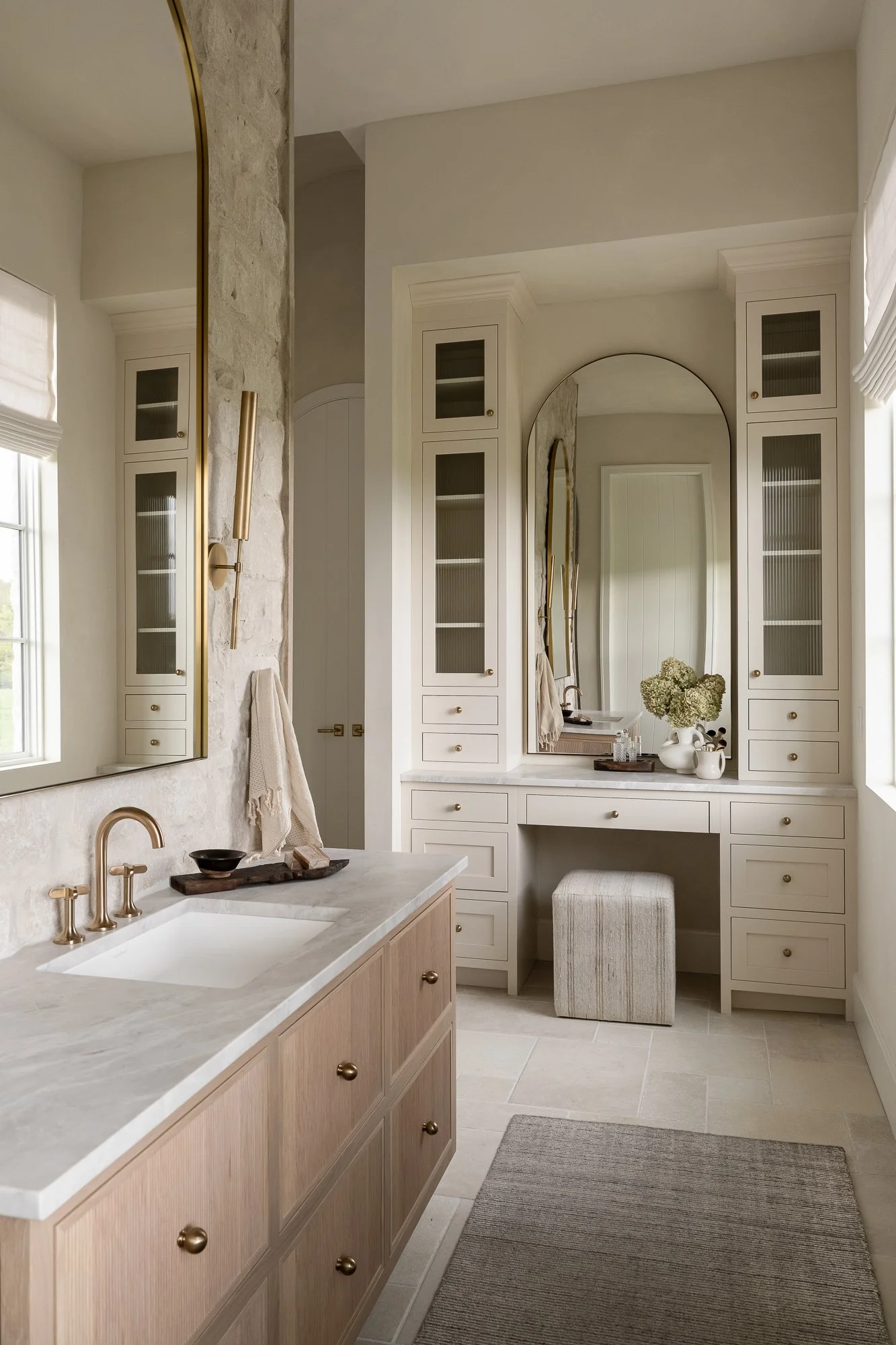 A bathroom with white cabinets, a large mirror, a white vanity with a floral arrangement, and a beige ottoman. Beige tiled flooring, a beige bath mat, and a window with white blinds are also visible.