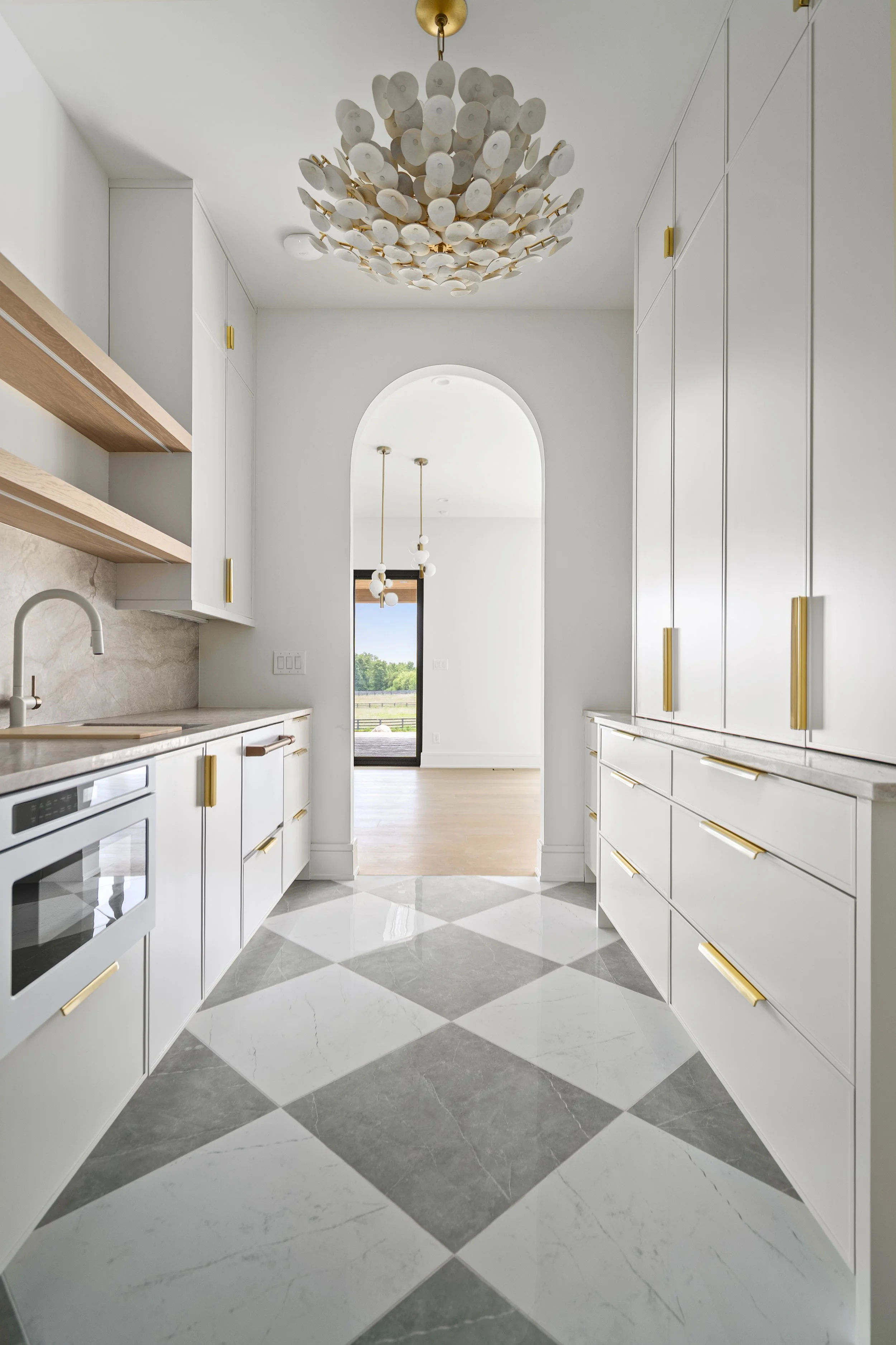Pantry with white cabinets gold handles, marble countertops, checkered tile floor, arched doorway leading outside, and modern chandeliers.