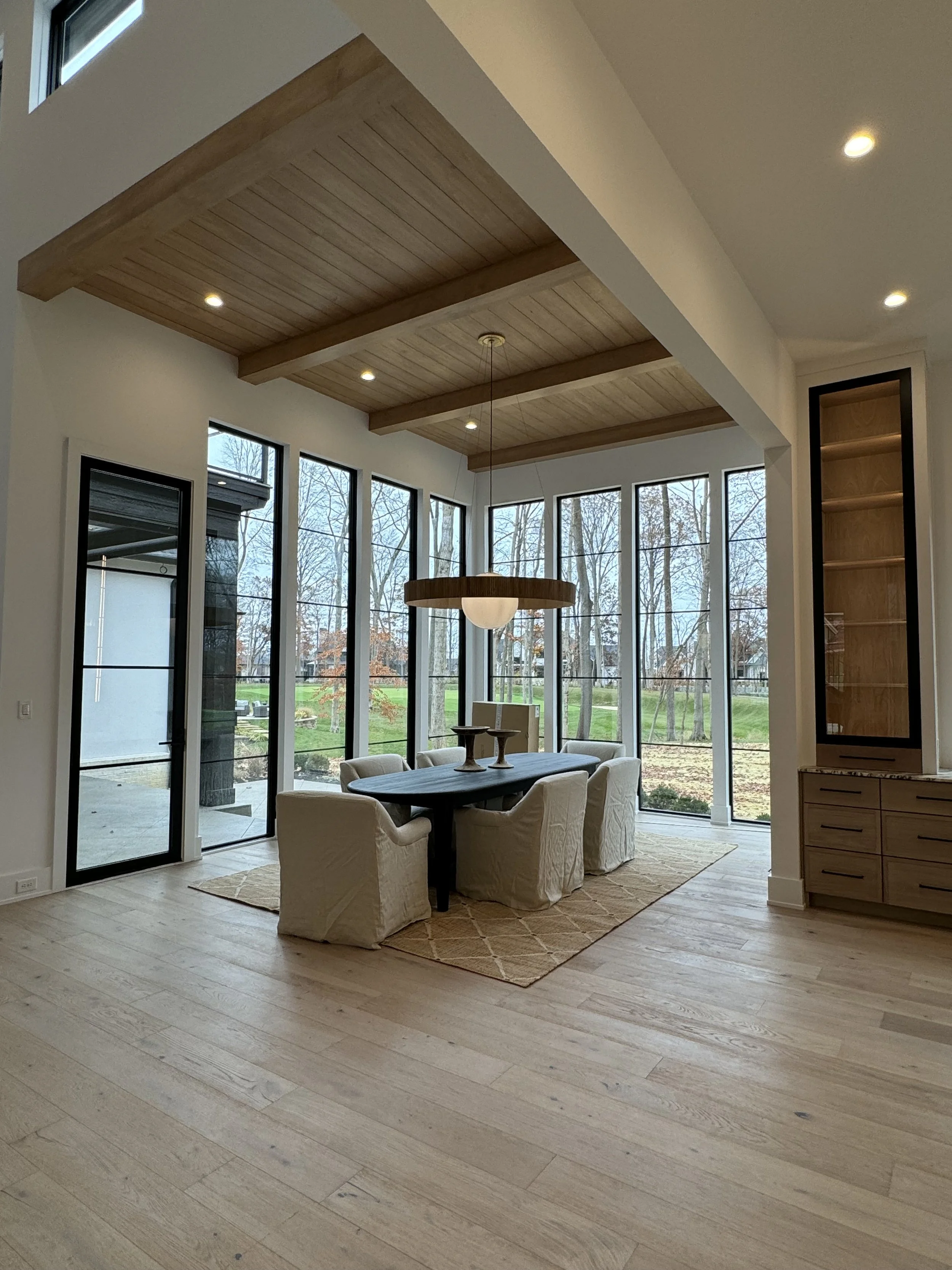 A bright dining area with large windows showing an outdoor scene of trees and a lawn. There is a wooden dining table with seven upholstered chairs, a hanging pendant light over the table, and built-in wooden shelves in the wall. The ceiling has woode