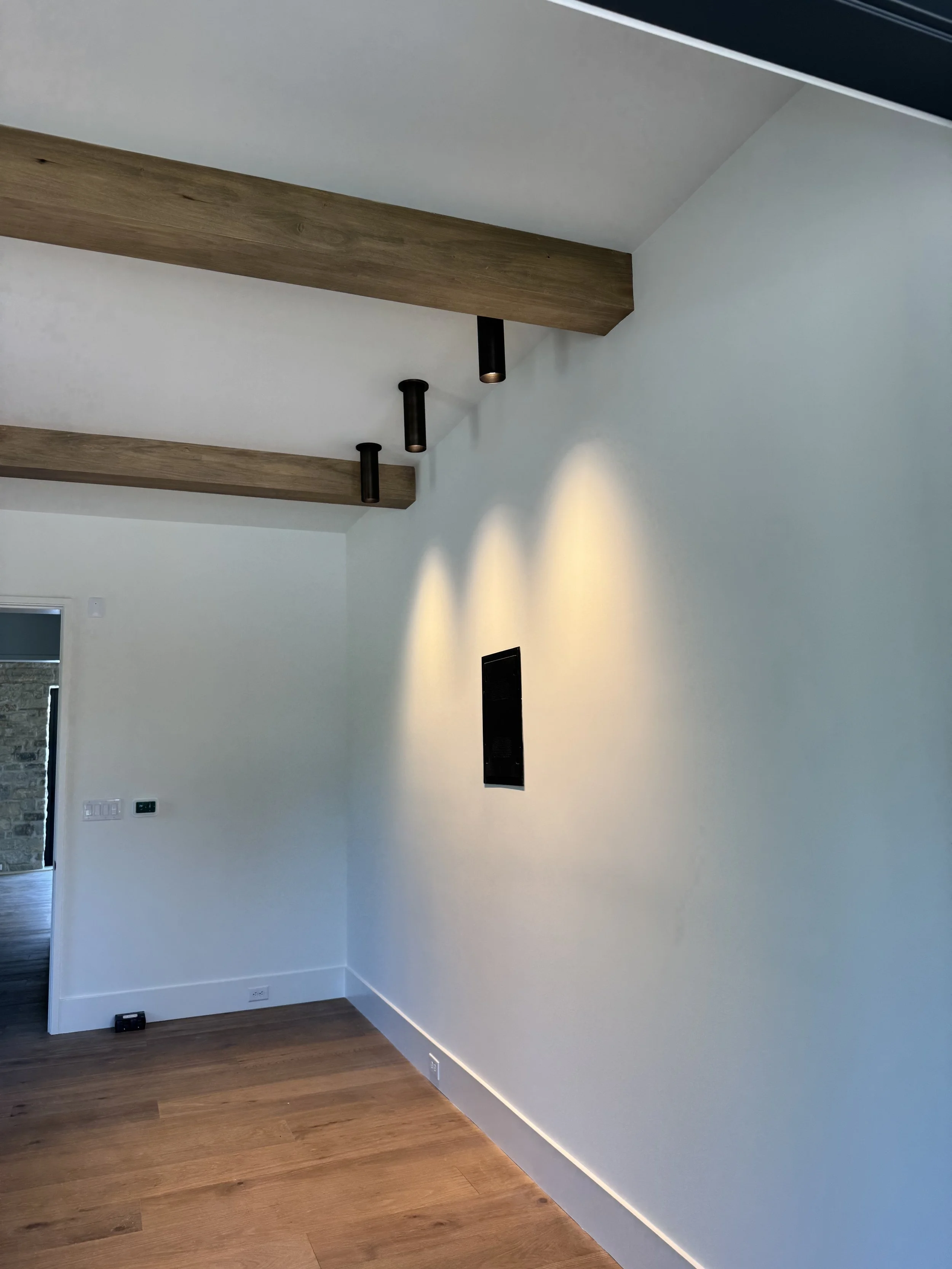 A minimalist room with wooden flooring, white walls, ceiling lights, and a corner with a wall vent and electrical outlets.