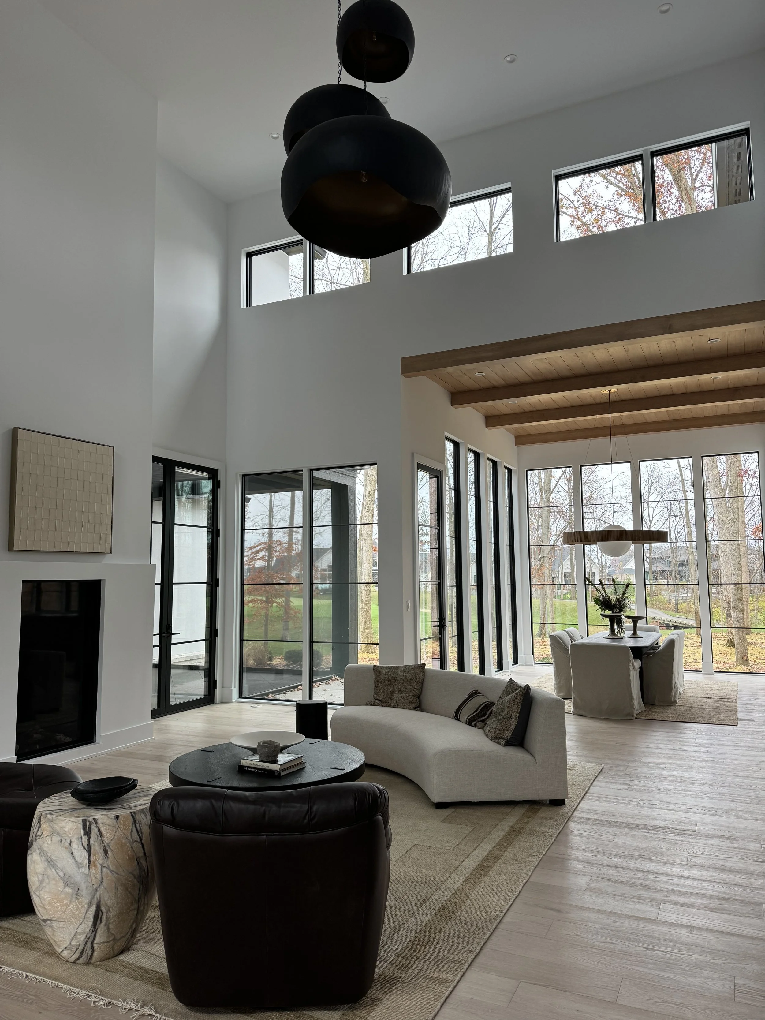Modern living room with floor-to-ceiling windows, a white sofa, black armchair, marble side table, and wooden beams on the ceiling. Outside trees are visible through the windows.