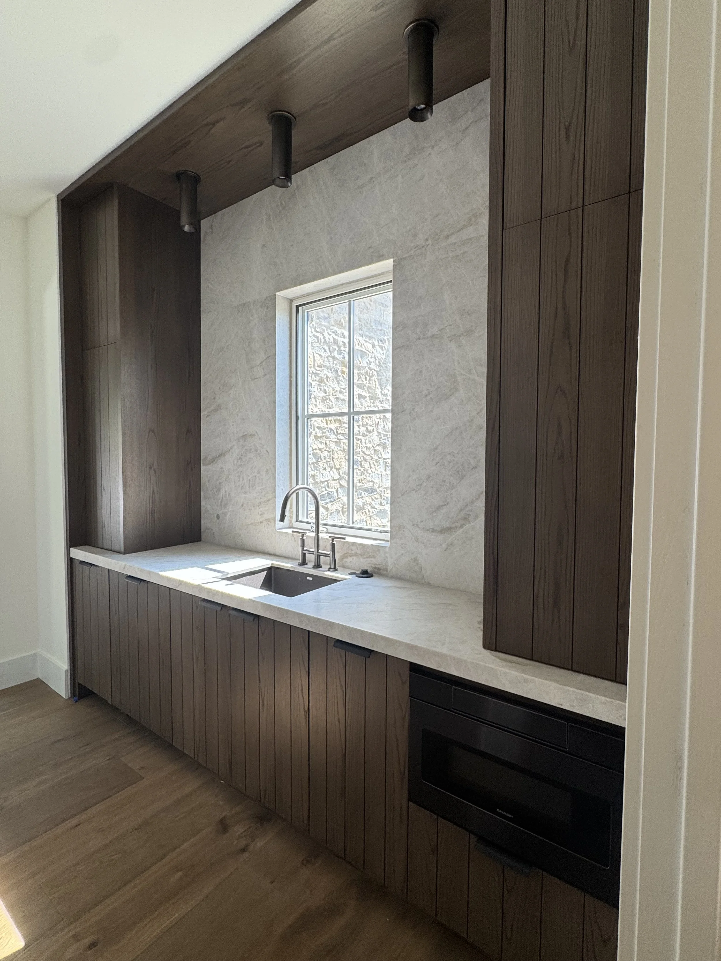 Modern kitchen with wooden cabinets, marble countertop, stainless steel sink, black oven, and a window showing a stone exterior wall.