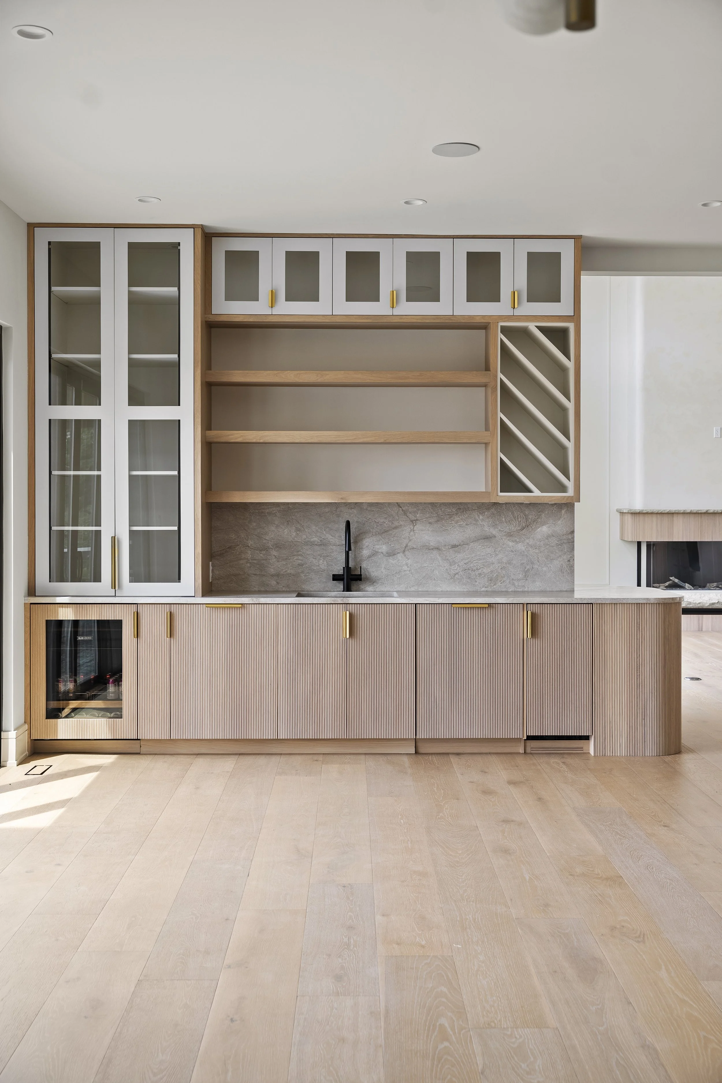 Modern kitchen with light wood cabinetry, black faucet, and a marble backsplash, with a large open shelving unit and glass-fronted cabinets, and beige hardwood flooring.