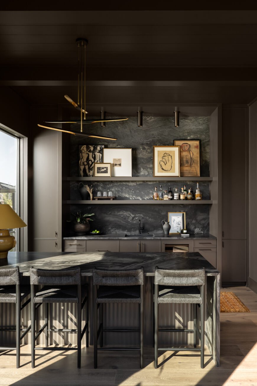 Modern kitchen with dark cabinetry, marble backsplash, open shelving with art and bottles, a dark marble island, and barstools with woven seats.