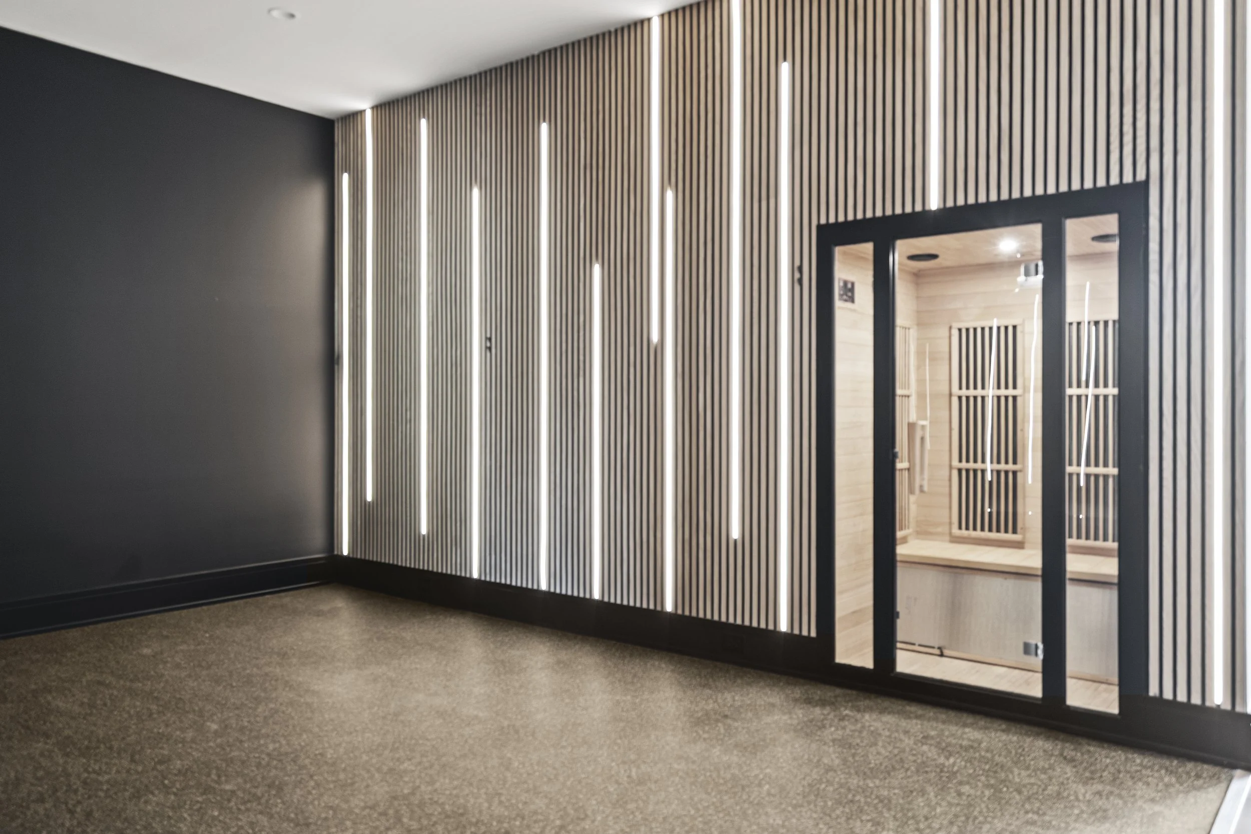 Modern gym with black and wood-paneled wall, vertical light strips, and an open doorway revealing a sauna with wood benches.