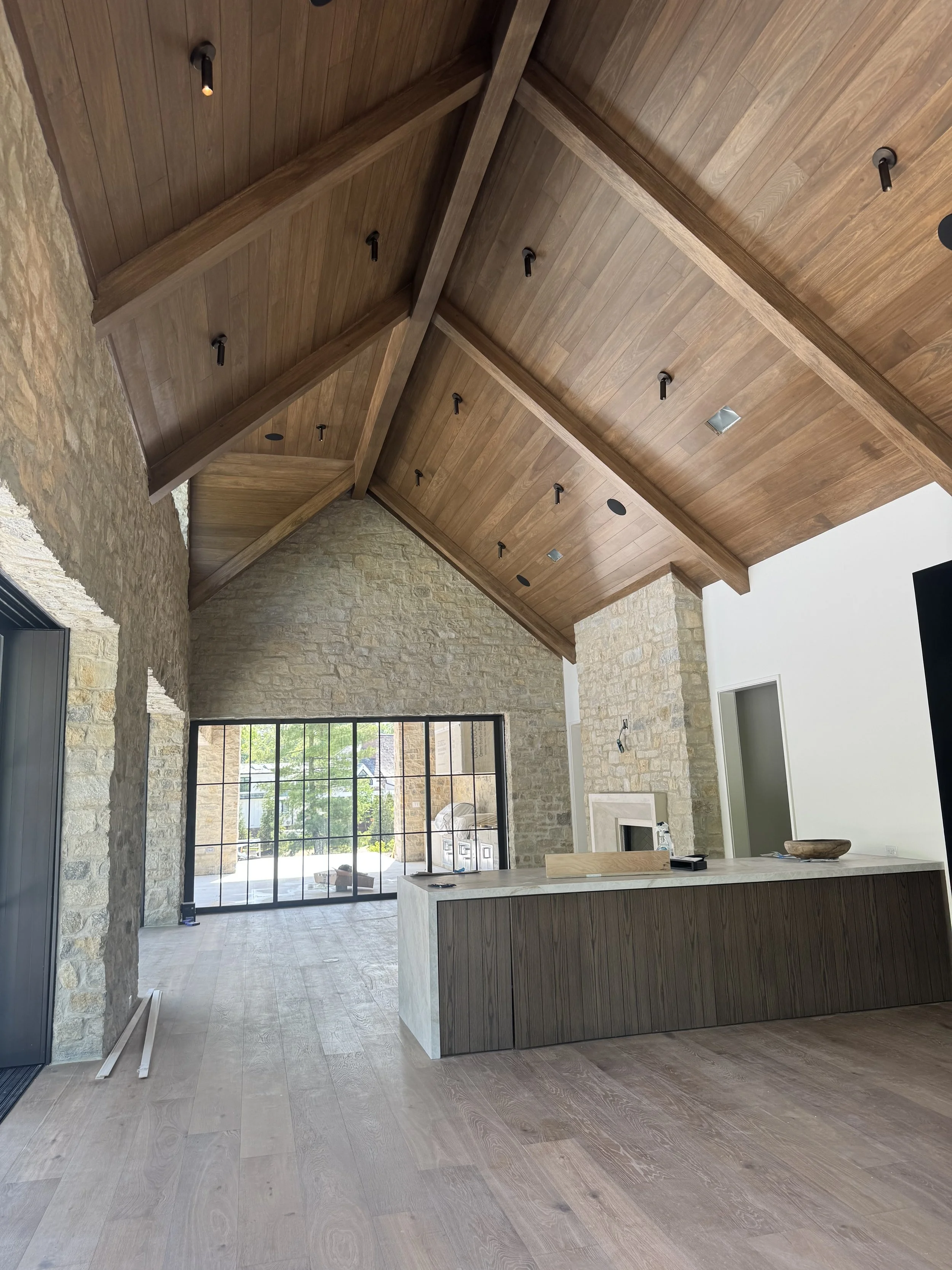 Interior of a modern home under construction with a large open living space, stone and white walls, wooden ceiling, large window, and an island with a marble top and wooden paneling.