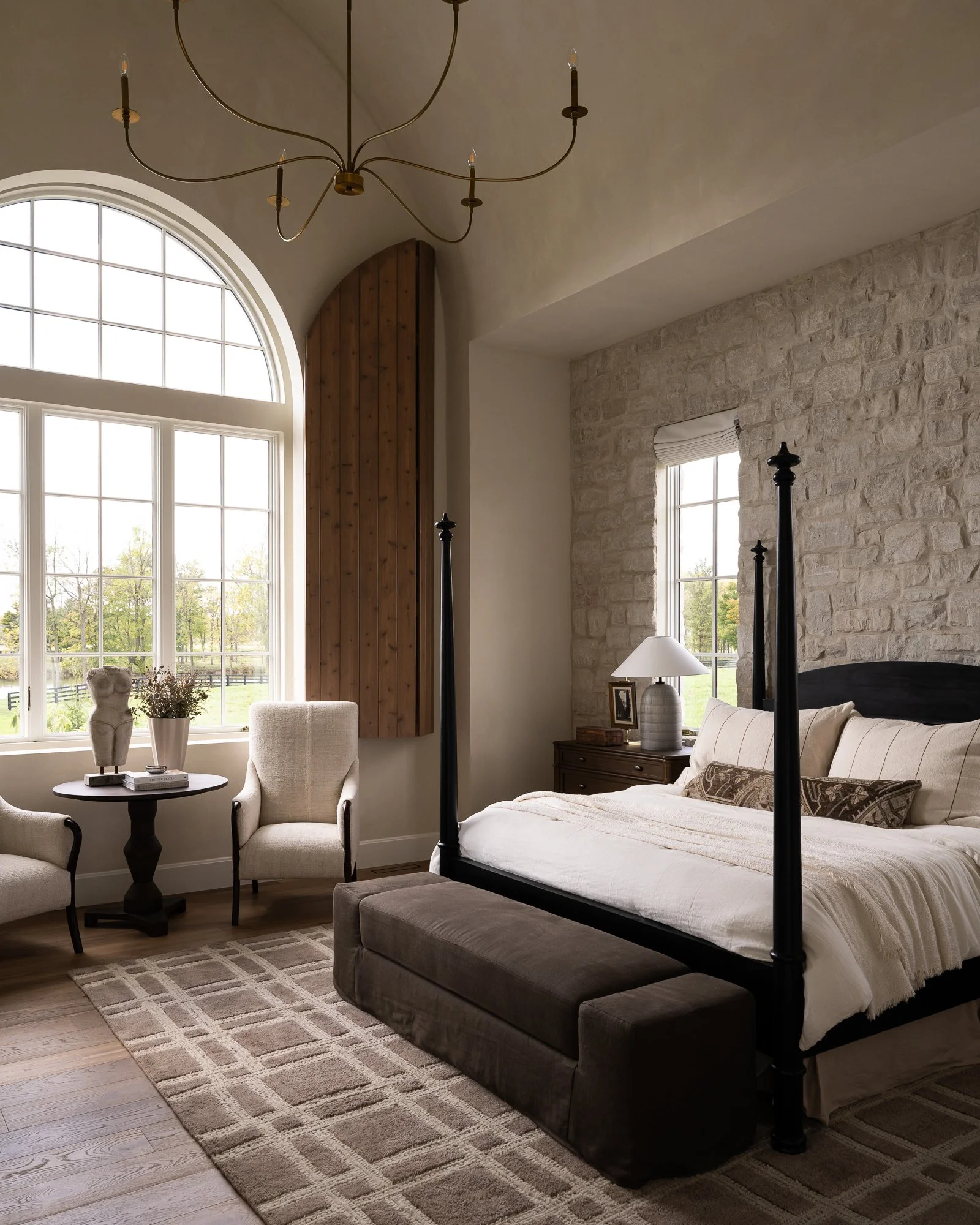 A bedroom with a large arched window, a stone wall, and a four-poster bed with white bedding and patterned pillows. There are two chairs, a small black table with a statue and books, a wooden nightstand with a lamp and framed photo, and a beige area 