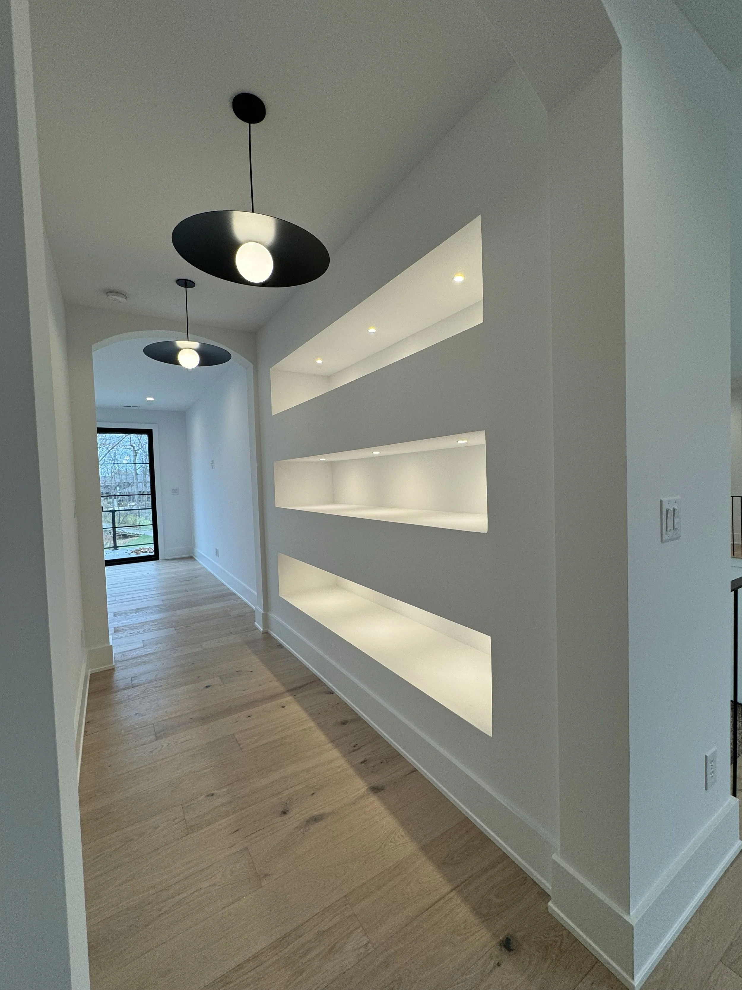 Modern living room with wooden floor, built-in white shelving, and two black pendant lights hanging from ceiling, with a window at the end showing trees outside.