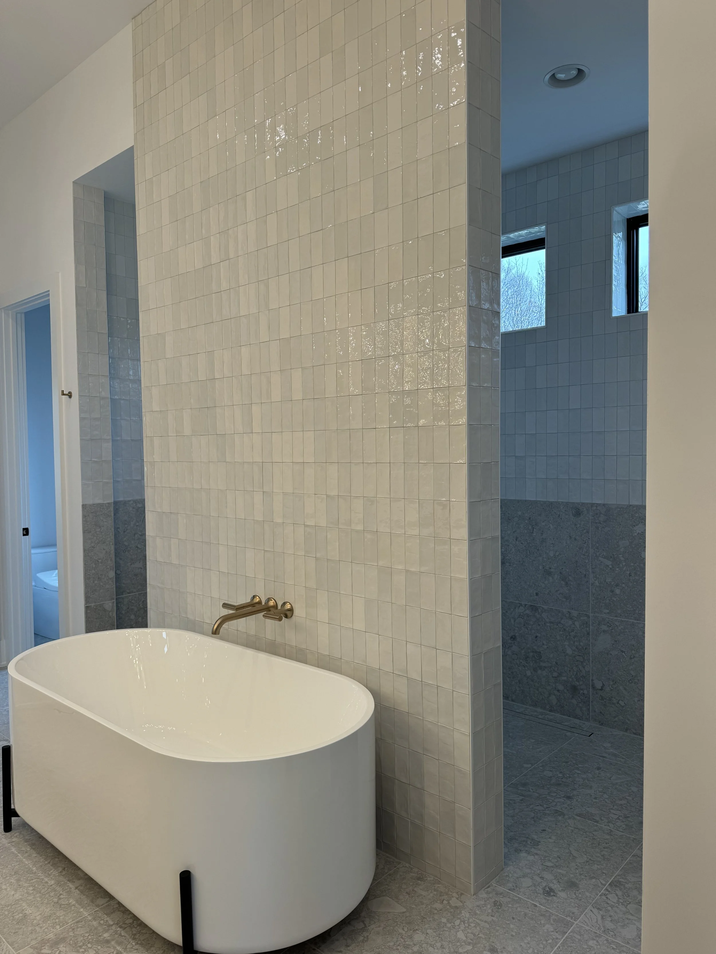 A modern bathroom featuring a white free-standing bathtub with a bronze faucet, adjacent to a wall covered in small square beige tiles. There are two large windows in the background allowing natural light, and the bathroom floor is finished with gray