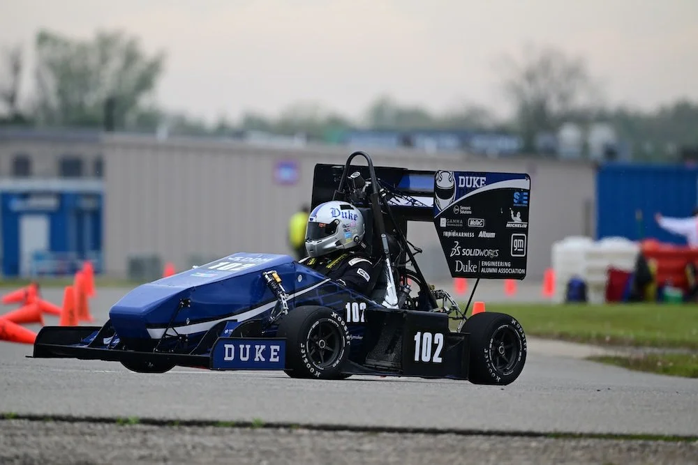 An open-wheel race car on a paved track, with a driver wearing a helmet inside the cockpit and a large rear wing featuring sponsorship logos. DDM Manufacturing Winston-Salem. motorsports machining background NASCAR