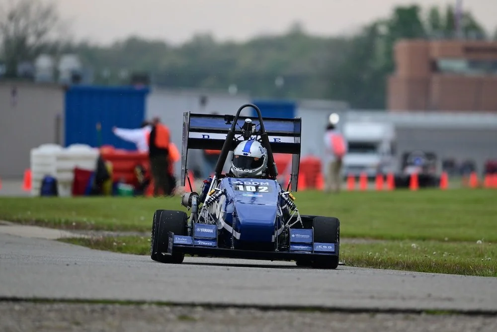 A blue Formula race car on a track during a race, with pit crew and cones in the background. motorsports machining background NASCAR