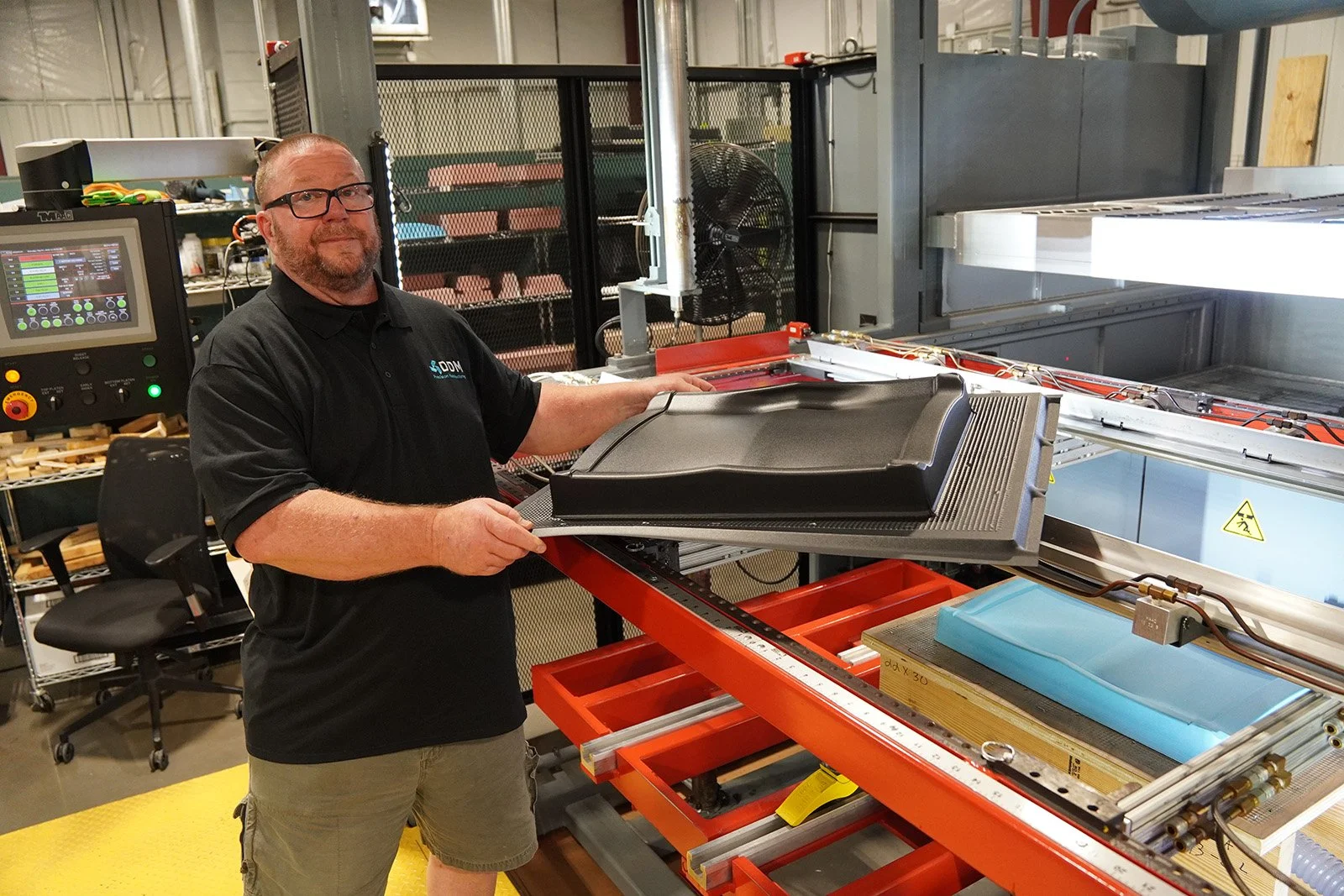 A man wearing glasses and a black polo shirt standing in a factory environment, holding a large black plastic or rubber part over a worktable with machinery, equipment, and metal structures in the background. aerospace defense manufacturing company l