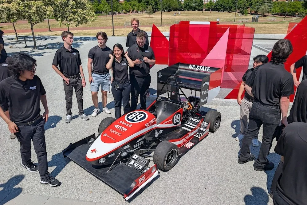 Group of people, mostly students, gathered around a red, black, and white race car with the number 89, on display outdoors with a red sculpture and trees in the background. DDM Manufacturing Winston-Salem.motorsports machining background NASCAR