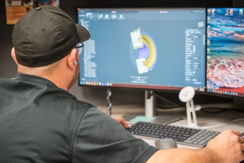 A person wearing a black cap working at a dual monitor computer setup. The left monitor displays a 3D modeling or design software.