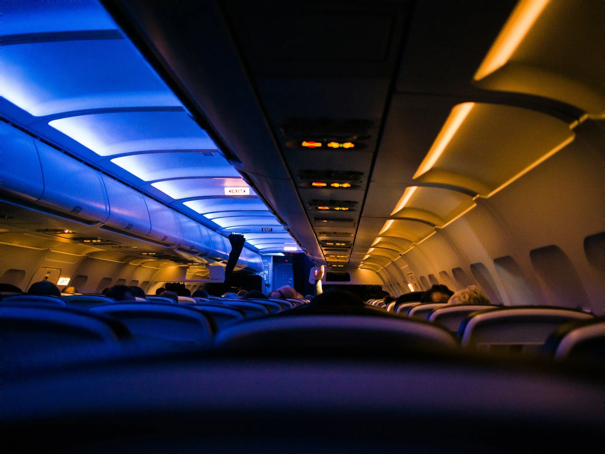 AIRCRAFT INTERIORS AND MRO - Inside an airplane cabin with illuminated blue and warm lighting along the ceiling, passengers seated, and an illuminated exit sign visible in the back.