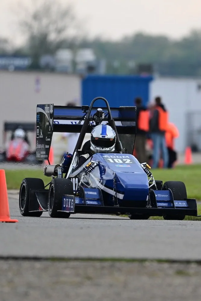 A blue Formula SAE race car driving on a track with orange cones, with spectators and a blue container in the background. DDM Manufacturing Winston-Salem.motorsports machining background NASCAR