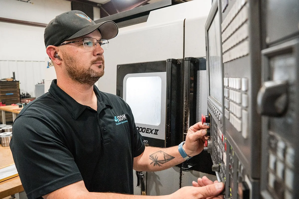 A man with a beard, glasses, a black cap, and a black polo shirt operating a CNC machine in a workshop. vacuum forming thermoforming aerospace parts