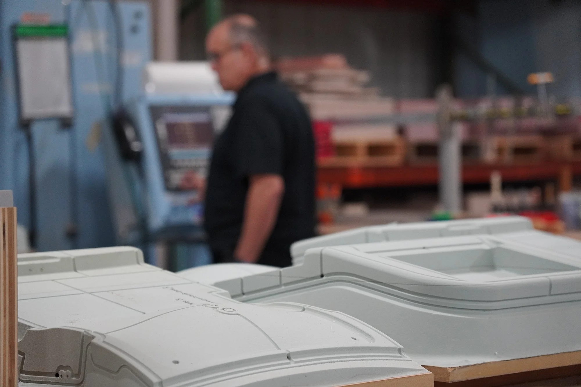 A person with glasses wearing a black shirt is working in a workshop or store, with shelves holding various items in the background and a large pile of white plastic objects in the foreground. single source machined and formed parts