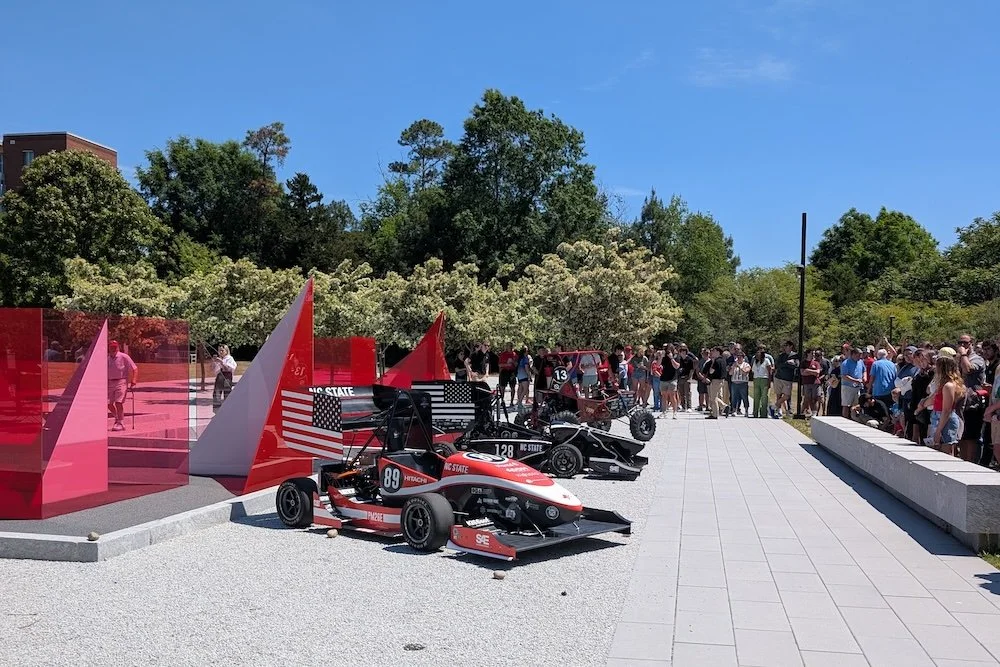 Race cars and off-road vehicles on display at an outdoor event with spectators watching. DDM Manufacturing Winston-Salem. motorsports machining background NASCAR