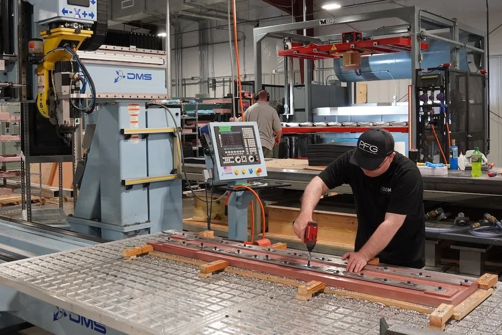 Man working on a metal piece with a power drill in an industrial manufacturing facility, with machinery and equipment in the background. motorsports machining background NASCAR