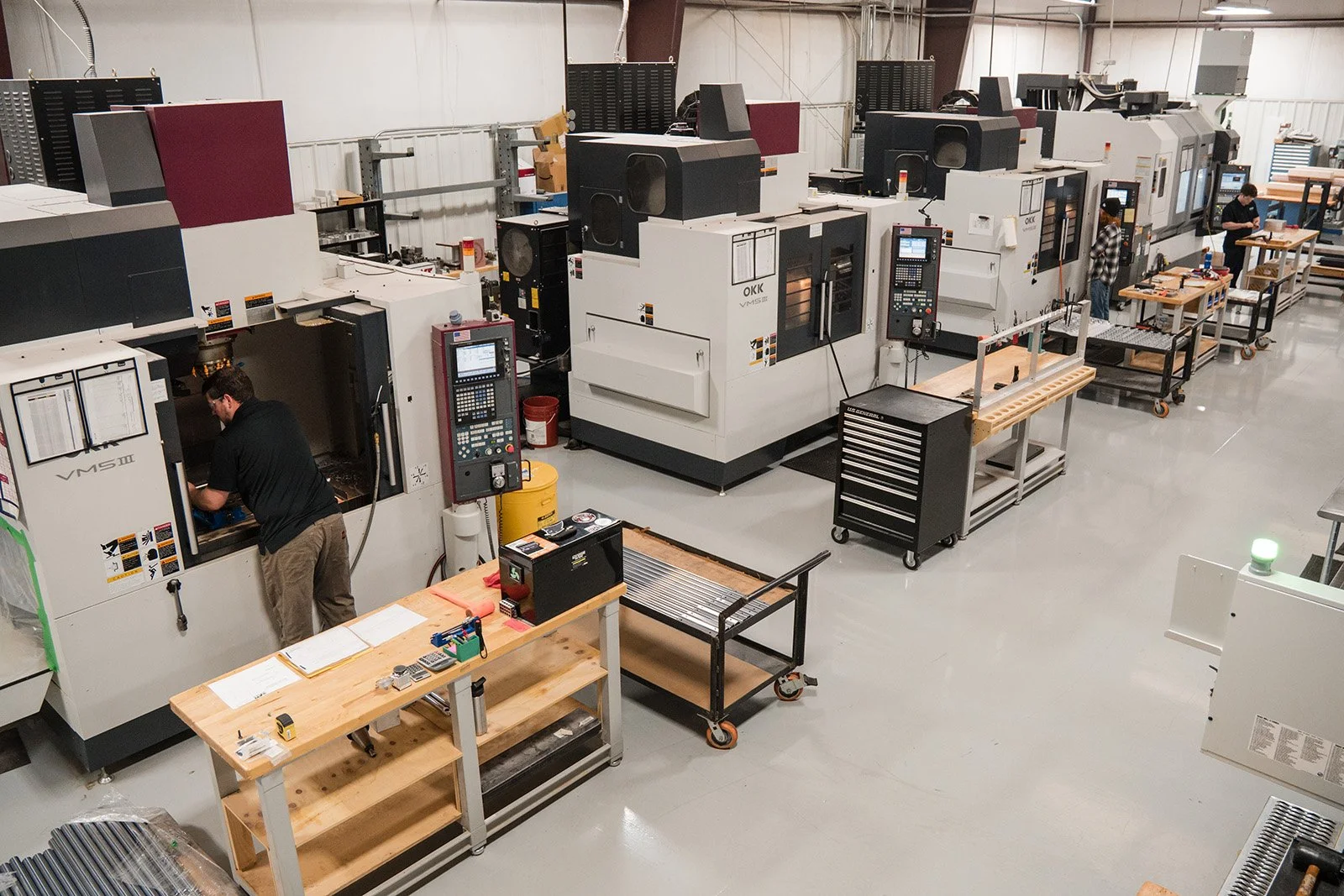 Industrial manufacturing factory with multiple CNC machines, workers operating equipment, and workbenches with tools and materials. 5-axis CNC milling turning aerospace defense