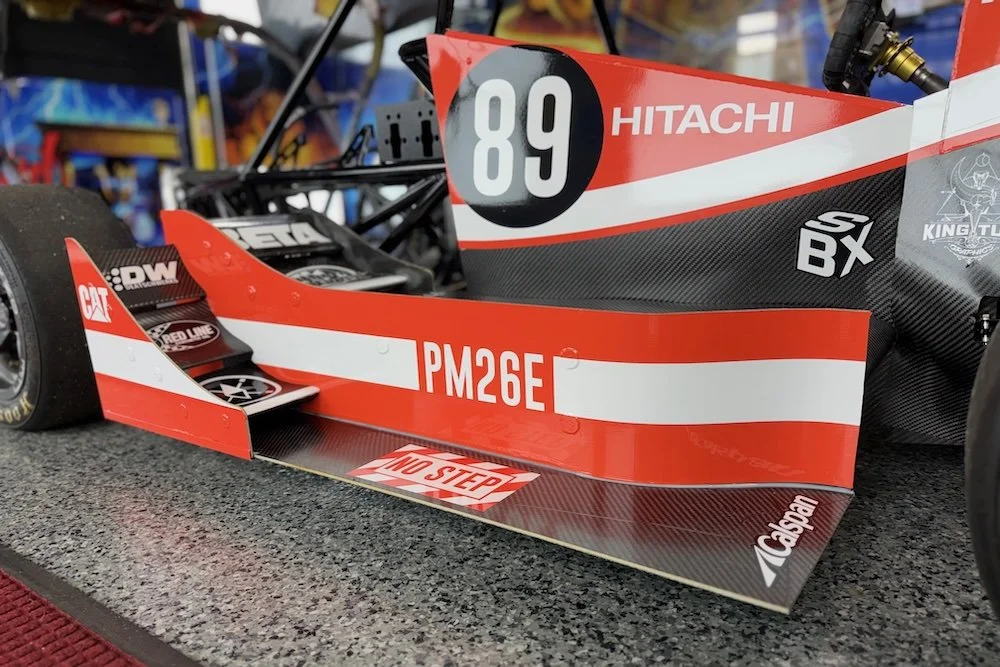 Close-up of a race car with red, white, and black graphics, displaying the number 89, sponsor logos like Hitachi, and a caution message on the front wing.motorsports machining background NASCAR