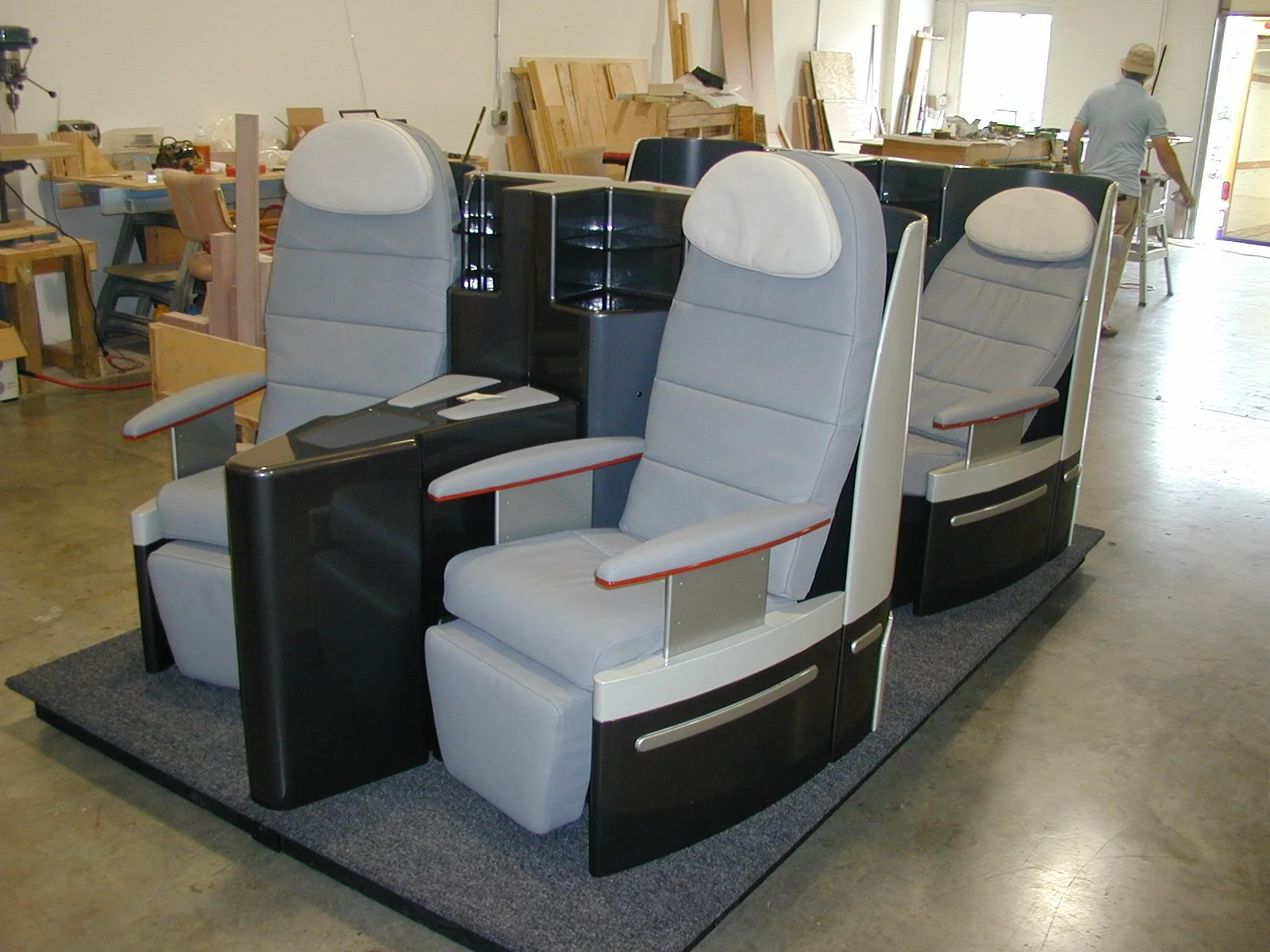 View of a row of replica Airbus first-class airplane seats in a workshop or showroom, with woodworking tools and materials visible in the background. manufacturing company Winston-Salem history