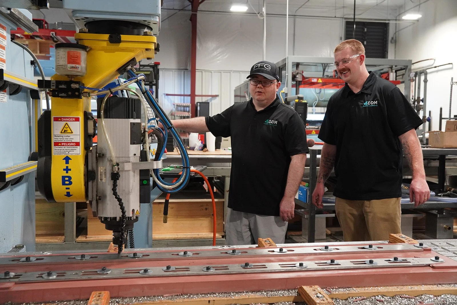 Two men working with a large industrial machine in a workshop. One is operating the machine while the other watches, both wearing black shirts with company logos.driven disciplined momentum manufacturing