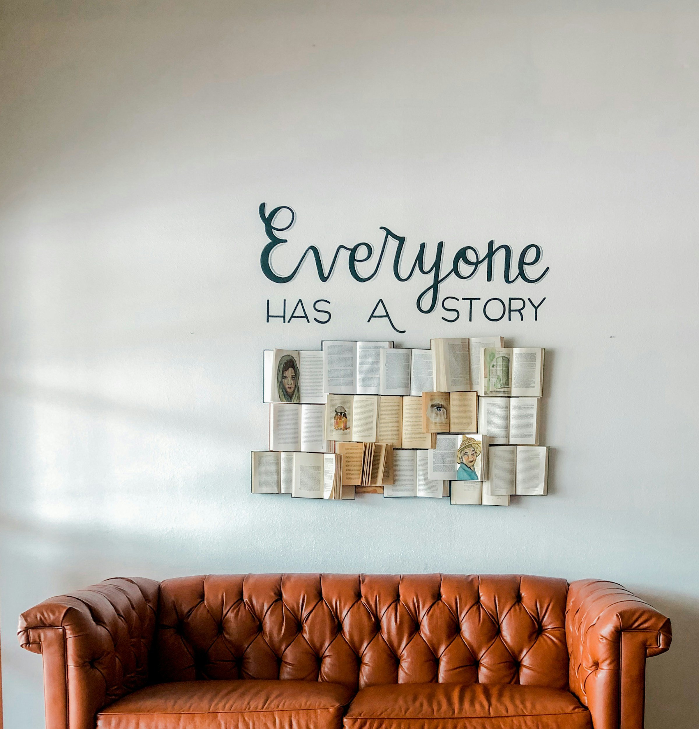 A brown leather tufted sofa in front of a white wall with a display of open books and illustrated portraits. Above the display, a quote reads: 'Everyone has a story'.