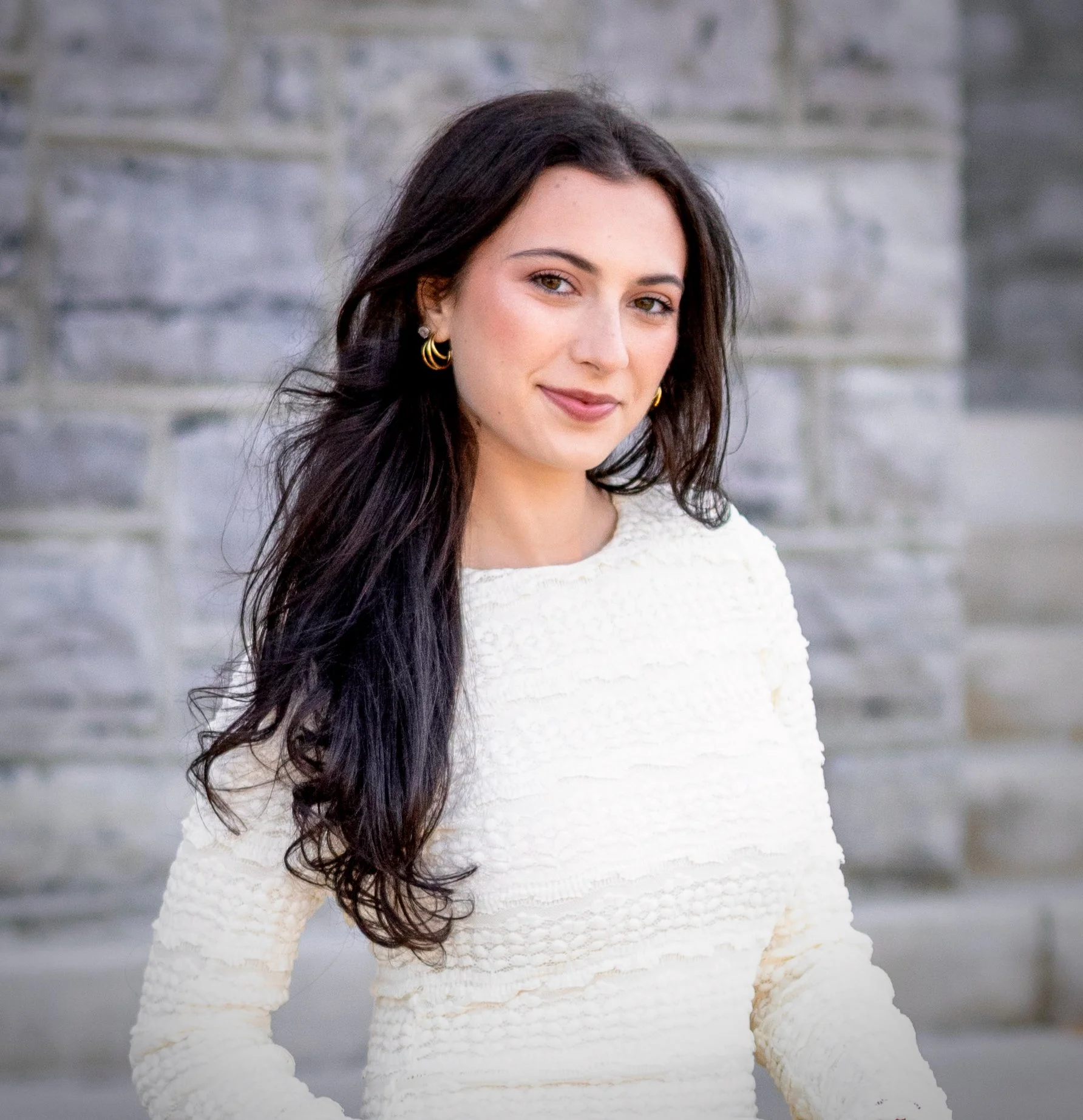A woman standing outdoors in front of a gray stone wall, wearing a white textured sweater and gold hoop earrings, with long dark hair and a slight smile.