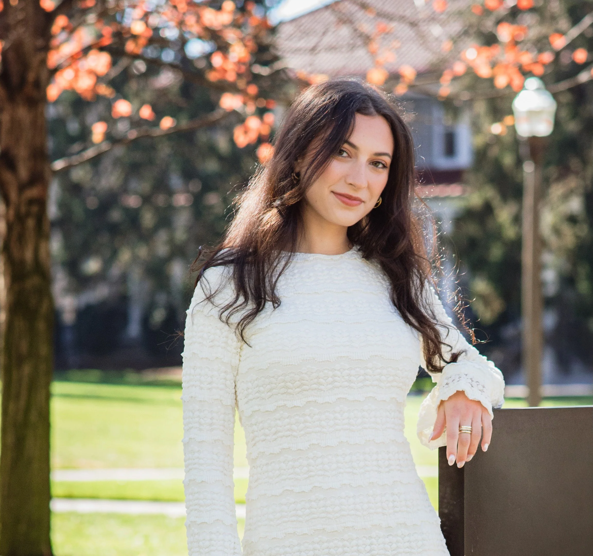 A woman with long brown hair, wearing a white textured dress, standing outdoors with a background of trees and warm sunlight.